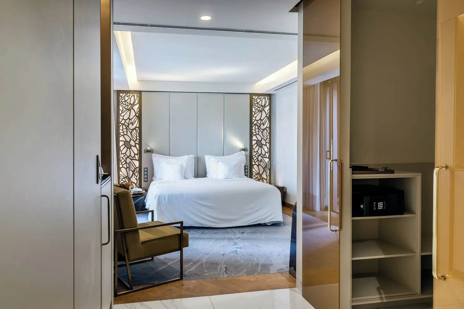 Avenue Room im Savoy Palace: ein Kingsize-Bett zwischen beleuchteten goldenen Gitterpaneelen, vom Eingang aus gesehen, vorbei an einem Sessel und einem Kleiderschrank mit Safe