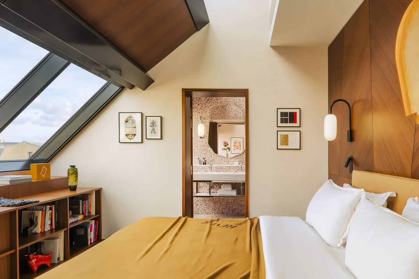 Suite Toits de Paris at Hôtel Madame Rêve: the bed beside a tall sloping atelier window, a low bookcase, and a doorway through to a mosaic-tiled bathroom with a long white basin