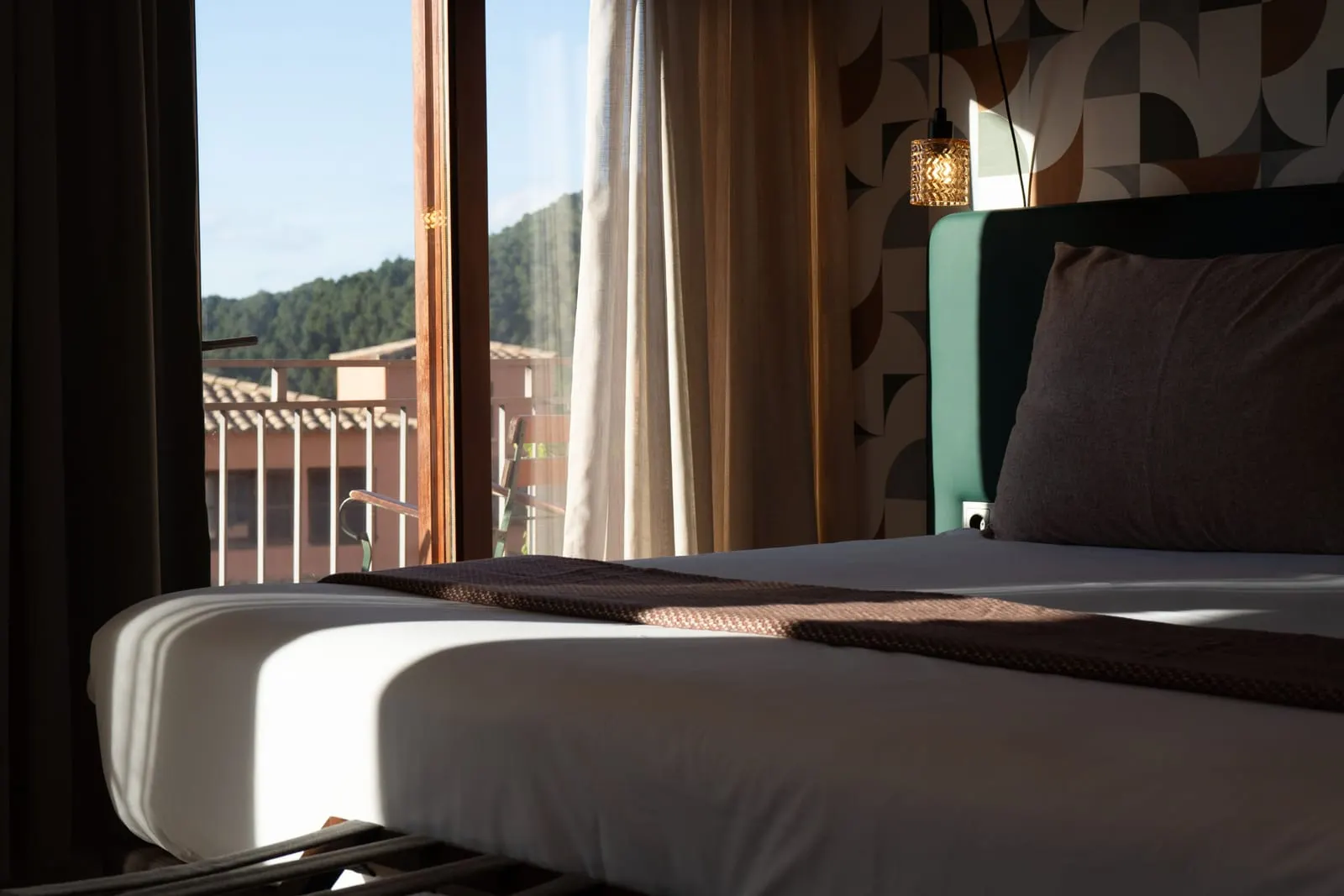 Sunset Hideaway at Maristel Hotel & Spa: a bed with a green headboard and patterned wall beside an open balcony door onto rooftops and wooded hills