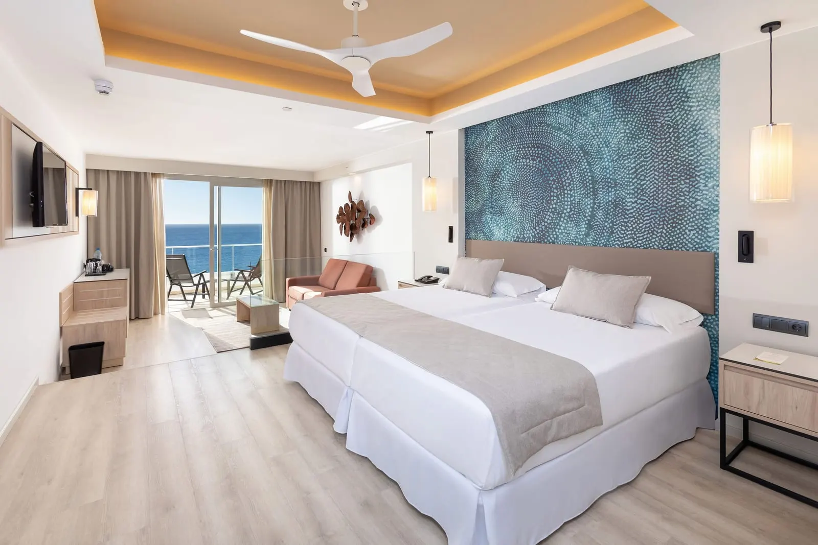 Double Room with sea view at Hotel Riu Palace Jandía: two beds pushed together under a patterned headboard wall, with a sofa beyond and a balcony door open to the sea