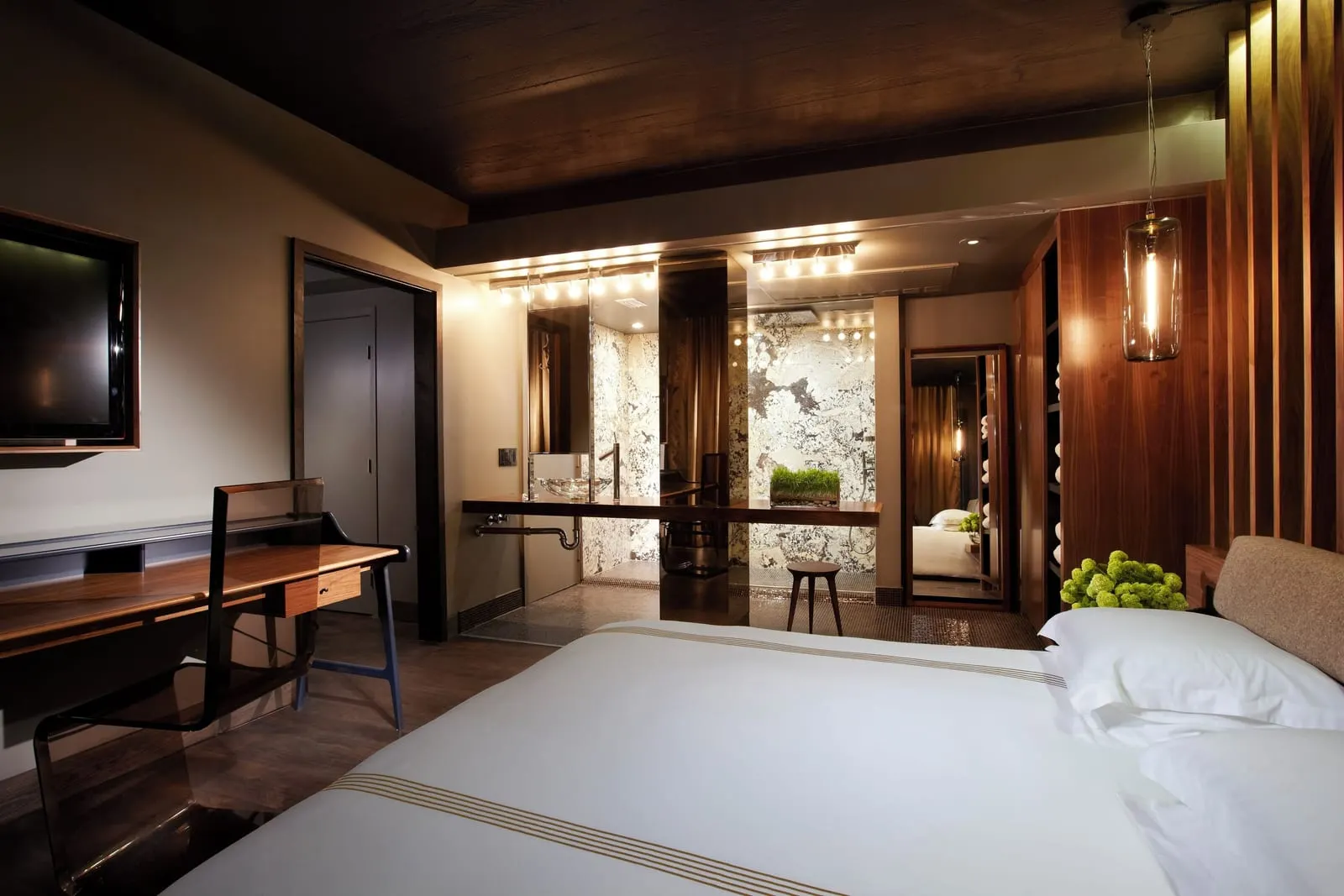 Tropicana King Suite im The Hollywood Roosevelt: ein Bett mit weißer Bettwäsche im Vordergrund, ein Schreibtisch und ein Wandfernseher, und dahinter ein Badezimmer mit Steinverkleidung und Waschtischleuchten