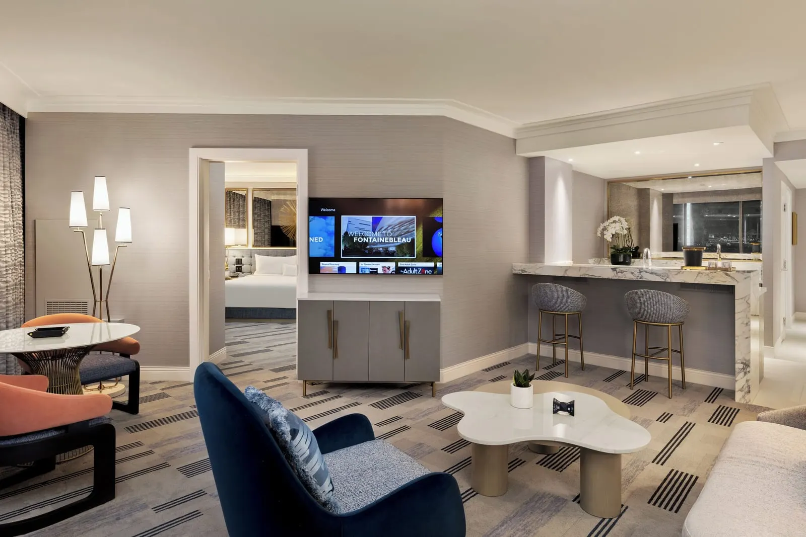 Noble Suite Pool View at Fontainebleau Las Vegas: the living room, with a navy armchair, a marble coffee table, a round table with two chairs, a marble bar with two stools and a doorway through to the king bedroom