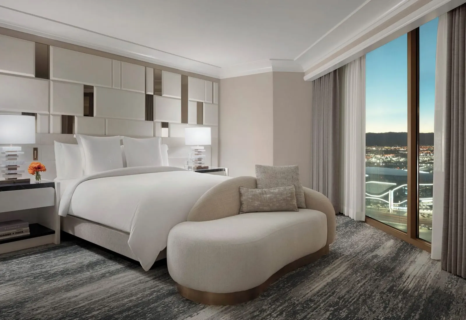 Strip View Presidential Suite at Four Seasons Hotel Las Vegas: a king bed against a panelled headboard wall, with a curved chaise at its foot and a tall window at dusk