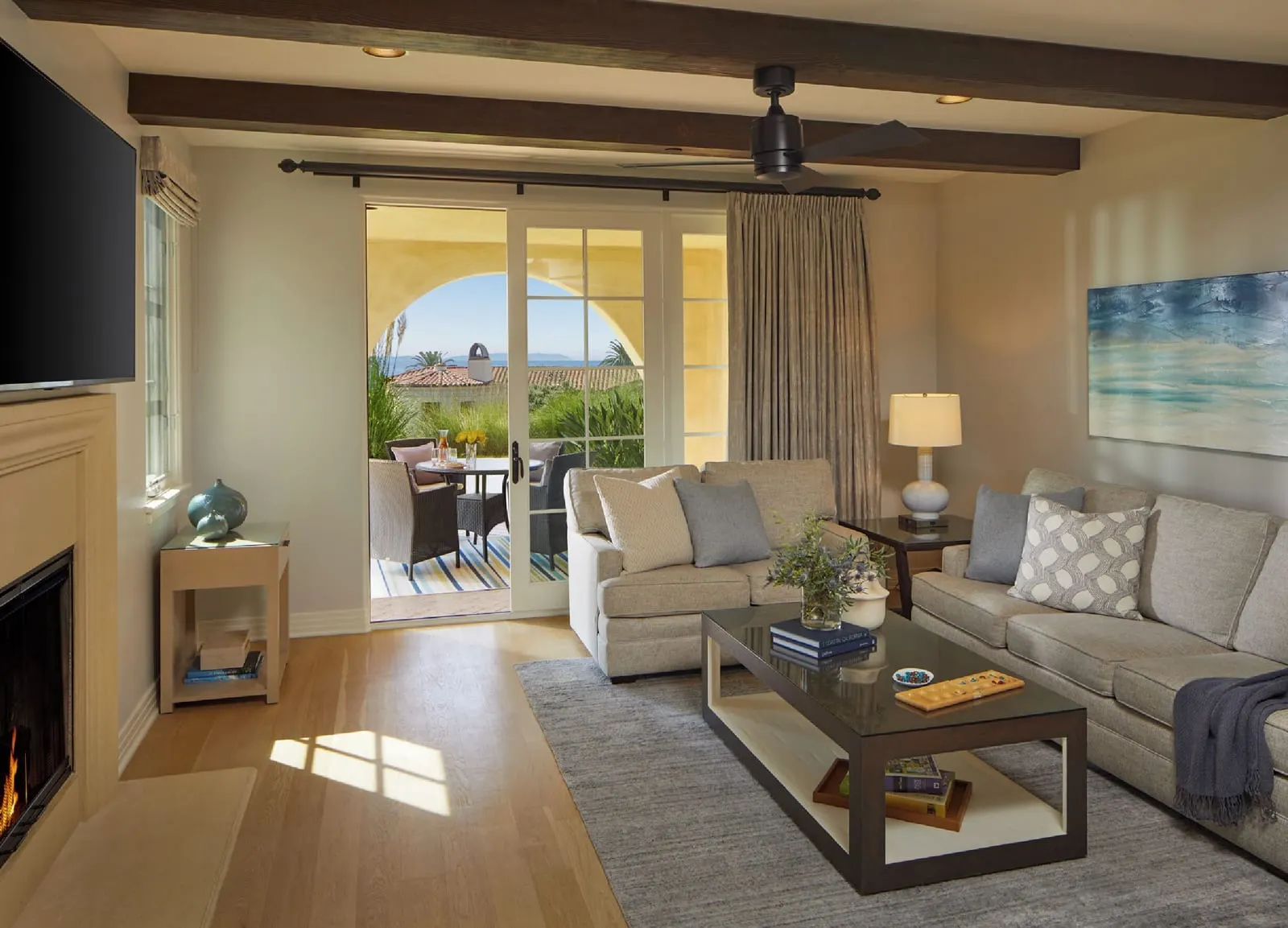 One-Bedroom Casita, Ocean View at Terranea Resort: a living room with a fireplace, a grey sofa, a wall-mounted television and glass doors to a covered terrace, under a beamed ceiling