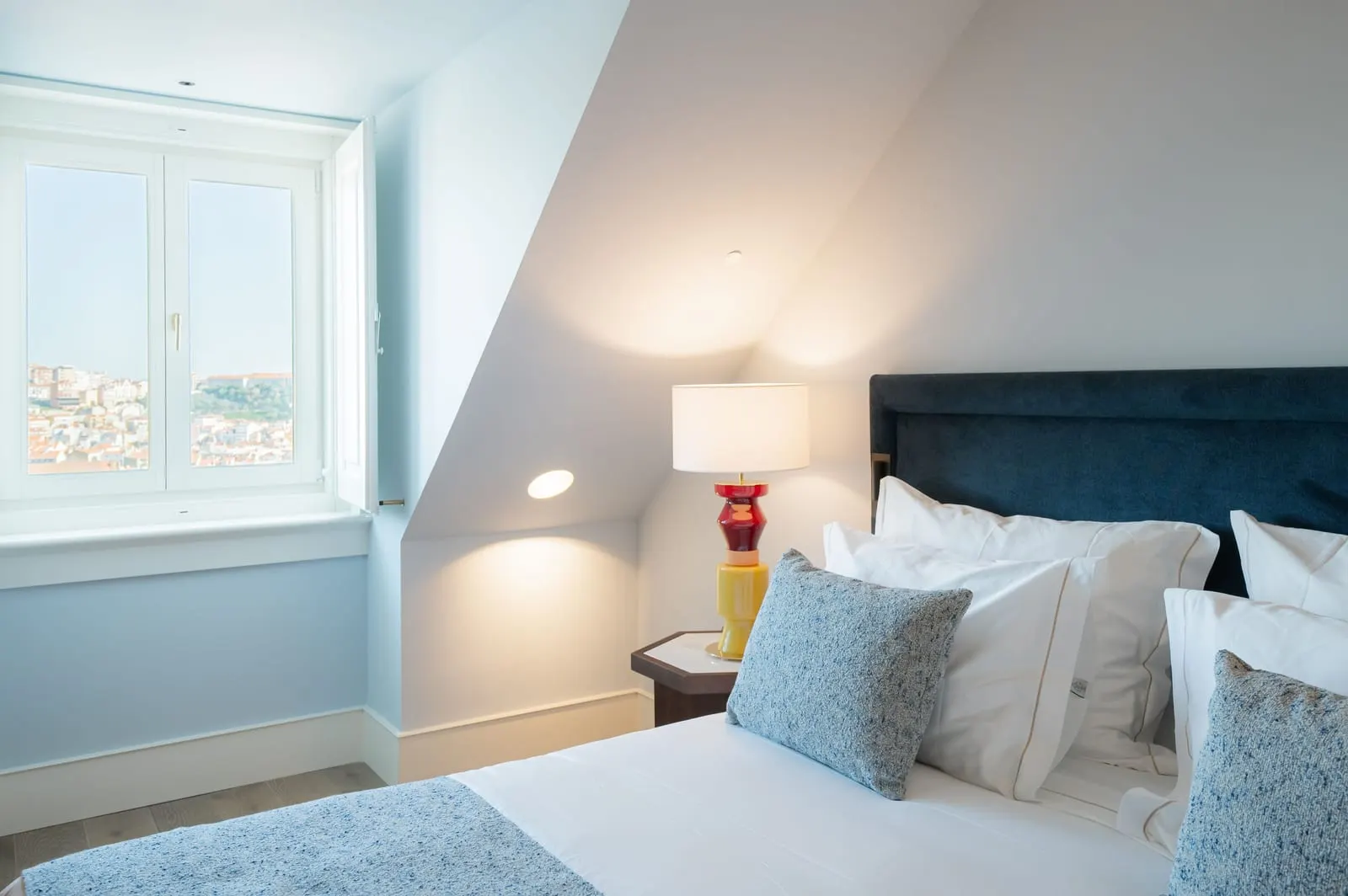 Suite at Palácio Ludovice: the bed with a navy headboard under a sloping ceiling, and a window looking across the city