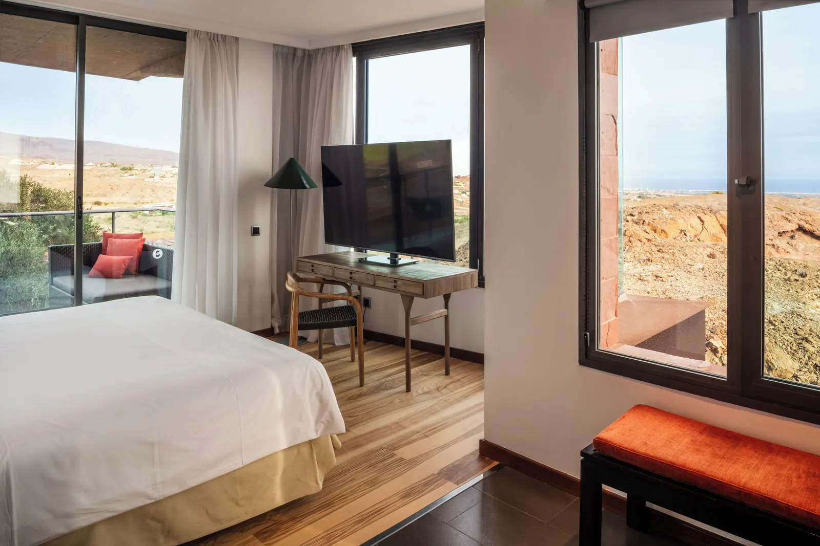 Deluxe Suite with Panoramic View at Salobre Hotel Resort & Serenity: the bedroom, with a bed, a desk with a television, an orange bench and windows on two sides