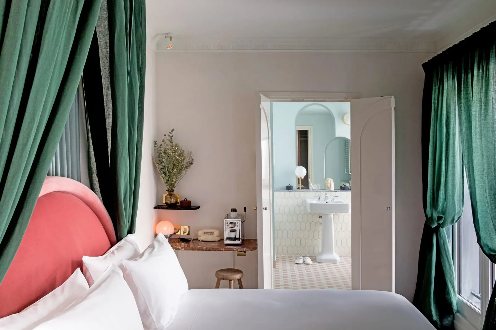Saint Fiacre at Grands Boulevards Experimental: a bed with a pink velvet headboard under dark green drapes, a marble shelf with an espresso machine and a rotary telephone, and double doors open onto a bathroom with a pedestal basin