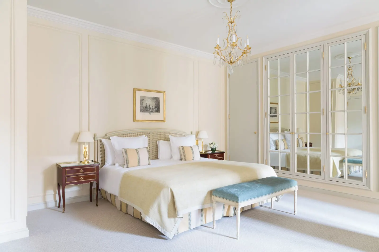 Junior Suite at Le Bristol Paris: a bed with a cream throw and a blue velvet bench under a crystal chandelier, beside mirrored wardrobe doors