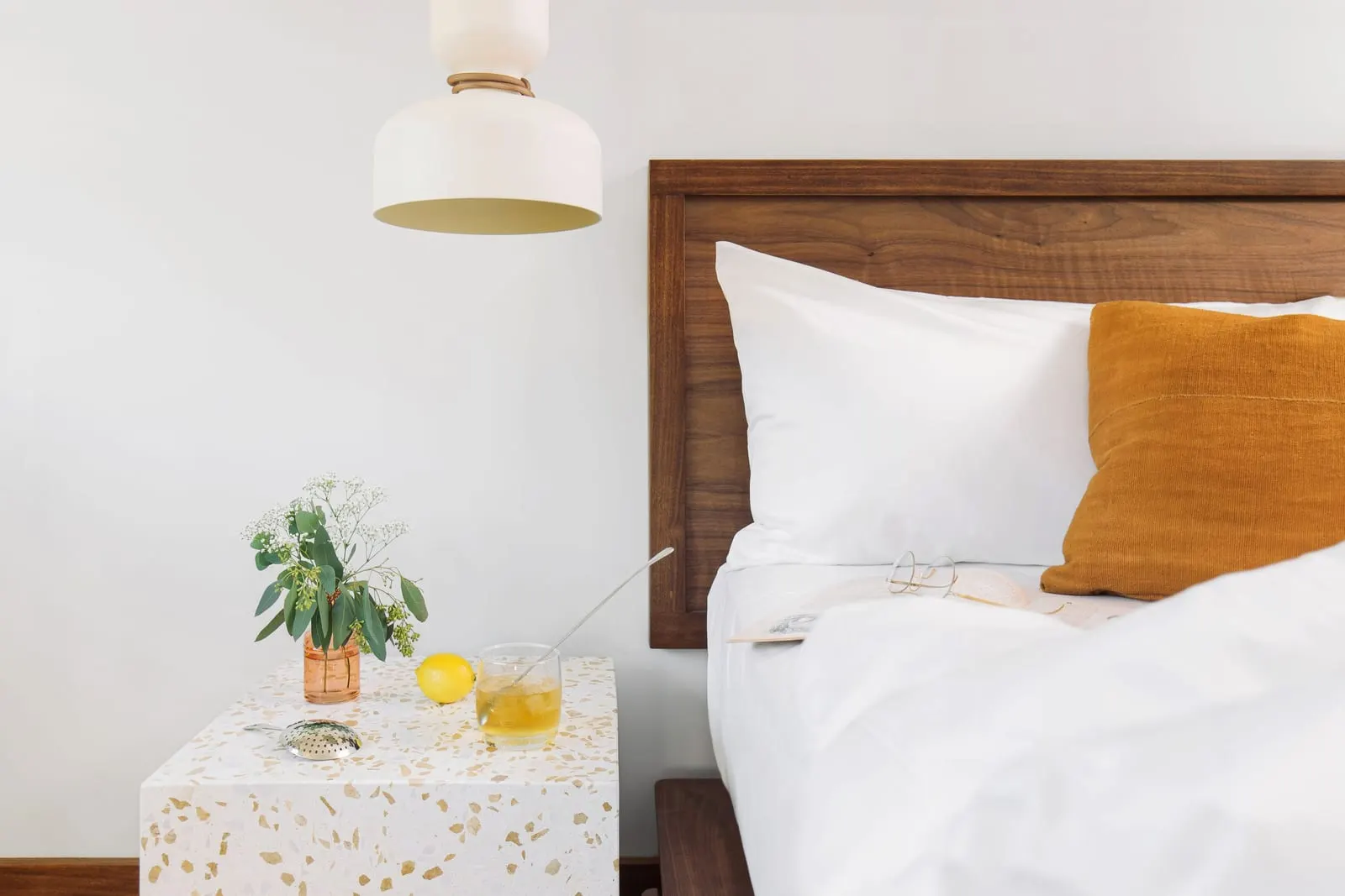 The Double Queen at Silver Lake Pool & Inn: a bed with white linen and a mustard cushion beside a terrazzo bedside table with fresh flowers, a lemon and a glass with a stirrer