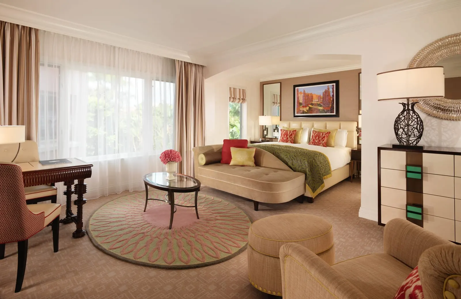 Deluxe Room at The Beverly Hills Hotel: a large room with a bed in an arched alcove, a tan chaise at its foot, a round patterned rug and a writing desk by sheer-curtained windows