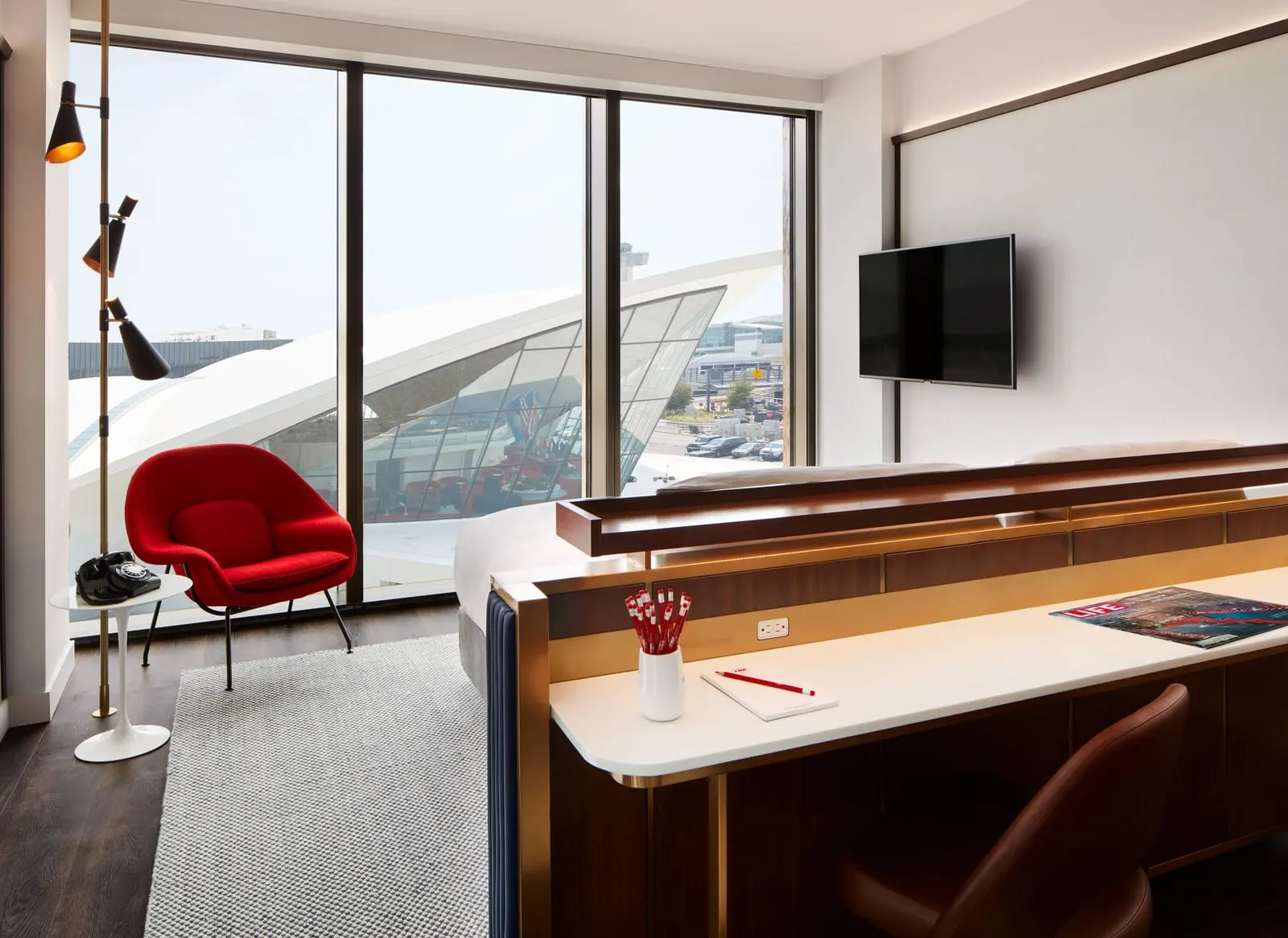 Deluxe King With Historic TWA View at TWA Hotel: floor-to-ceiling windows framing the white curved roof of the TWA Flight Center, with a red armchair and a rotary phone by the glass and the bed behind a long desk