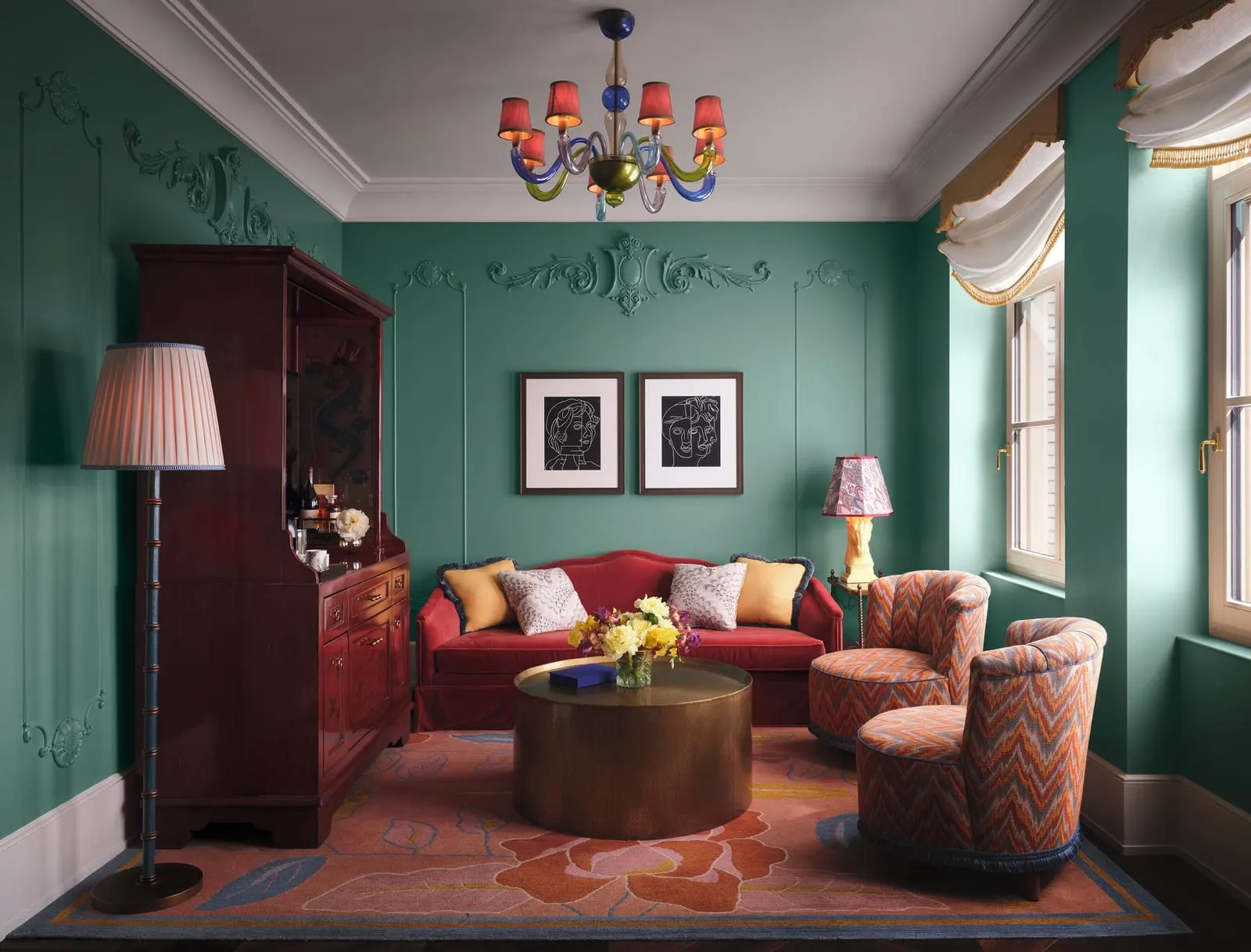Fifth Avenue Suite at The Fifth Avenue Hotel: a living room with green plaster moulded walls, a red sofa under two framed drawings, two patterned armchairs, a round brass table and a coloured glass chandelier