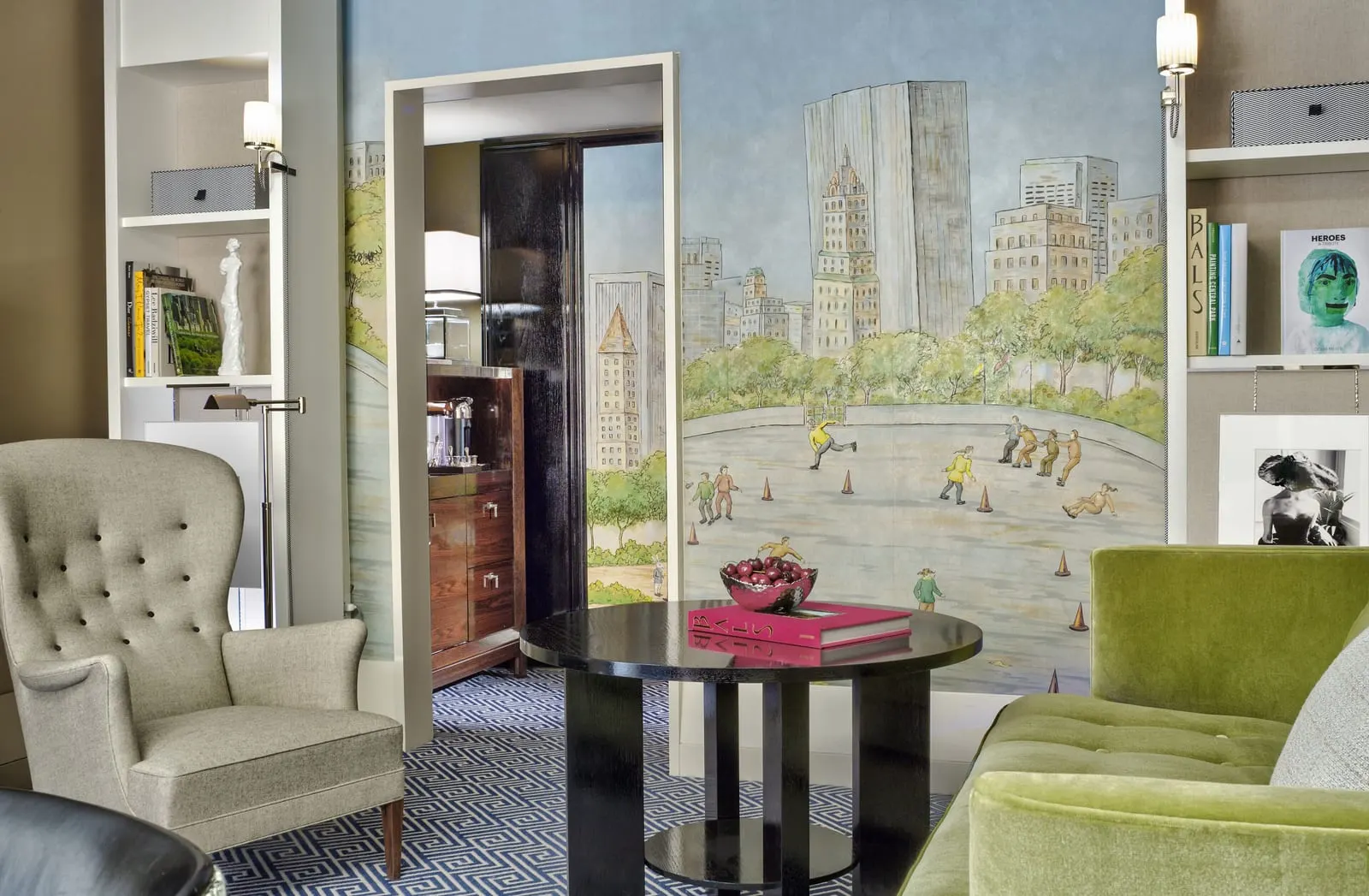 Premier Suite at The Carlyle: a living room with a green velvet sofa and a wing armchair in front of a painted mural of skaters, with a doorway beyond