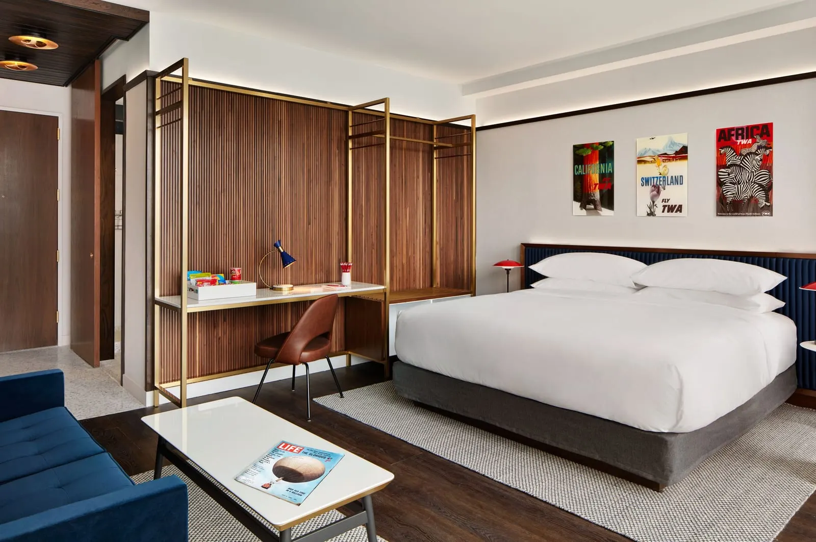 Executive King Suite at TWA Hotel: a king bed under three framed TWA posters, a desk in a brass-coloured frame against a walnut slatted wall, and a blue sofa and coffee table in the foreground