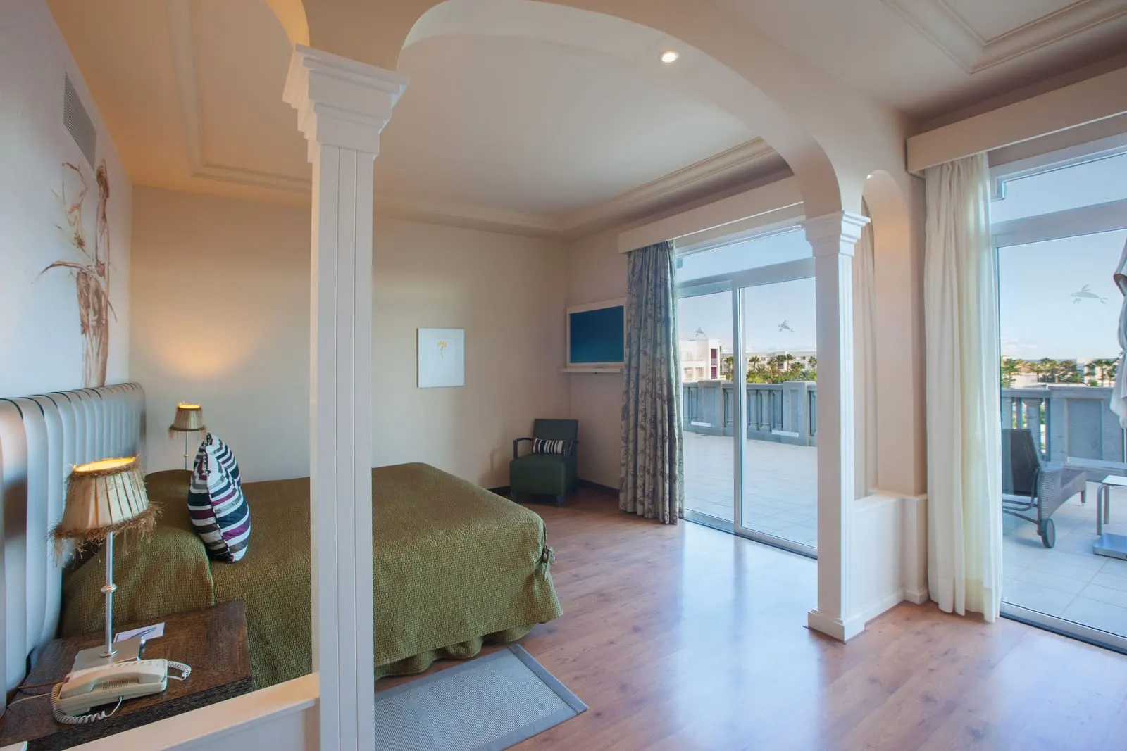 Senior Suite at Lopesan Villa del Conde Resort & Thalasso: a king bed with a padded headboard in an alcove framed by white columns and arches, beside sliding doors to a terrace with a sun lounger