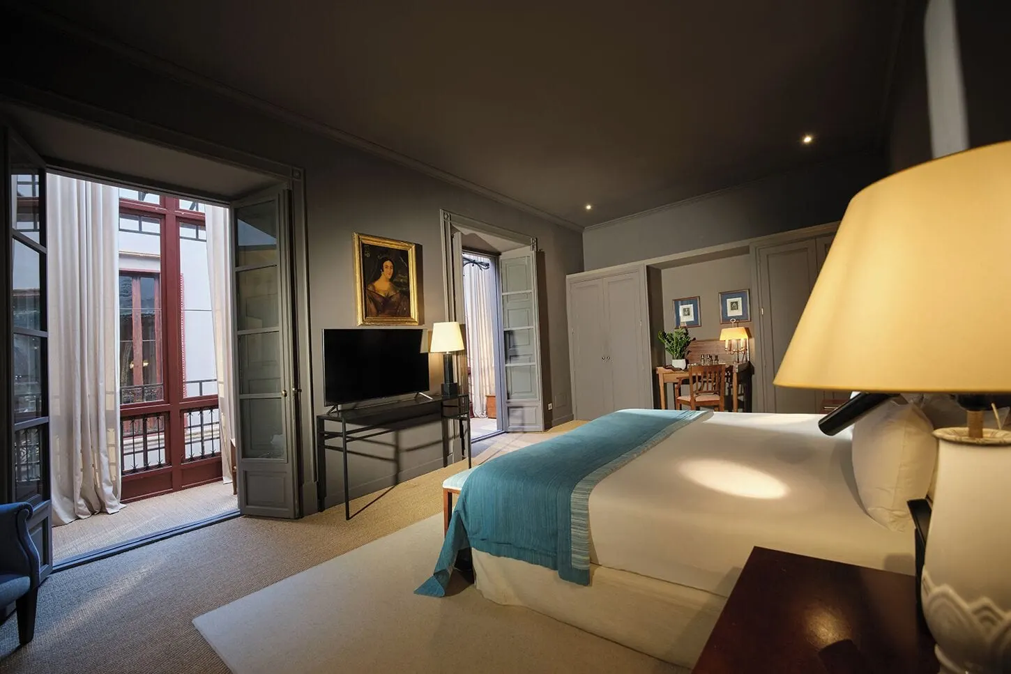 Signature Junior Suite at Can Cera: a bed with a teal throw, a television on a console, a framed portrait, a desk beyond and tall open doors with an iron balcony rail