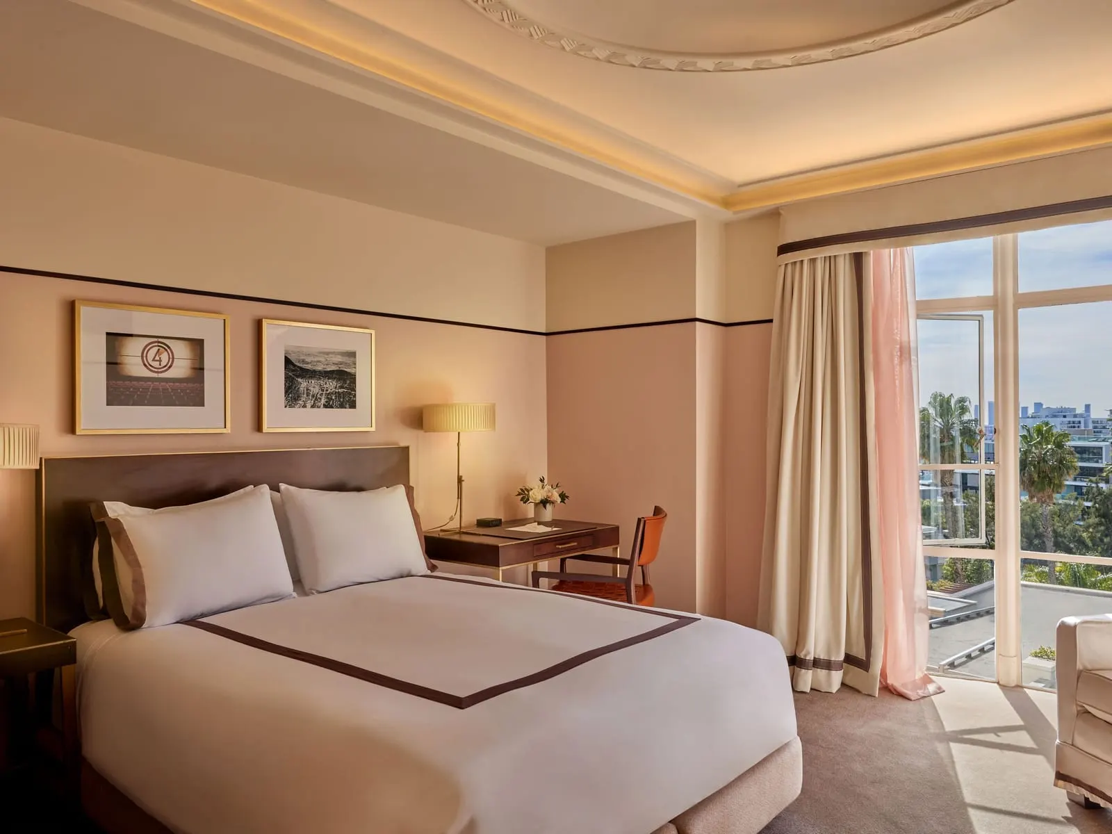 Superior Queen at Sunset Tower Hotel: a queen bed with a dark headboard, a writing desk and leather chair, framed prints and a tall window with cream and pink curtains