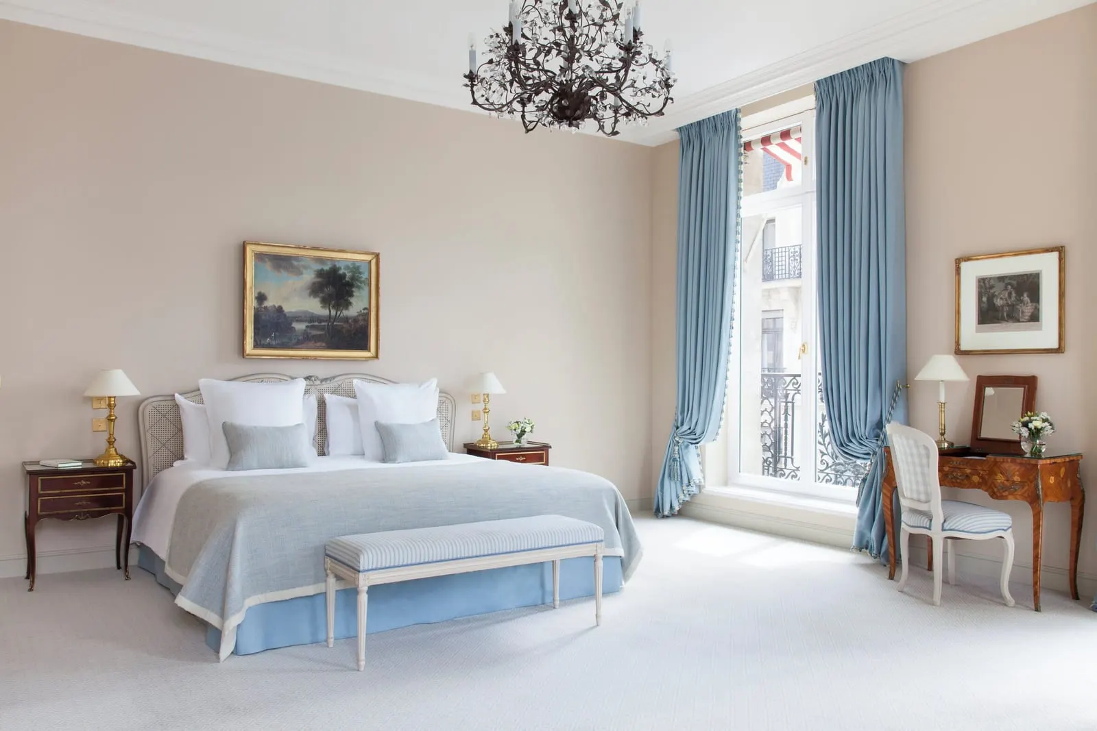 Suite Présidentielle at Le Bristol Paris: a bed with a pale blue cover and a bench under a wrought-iron chandelier, beside tall windows hung with blue curtains
