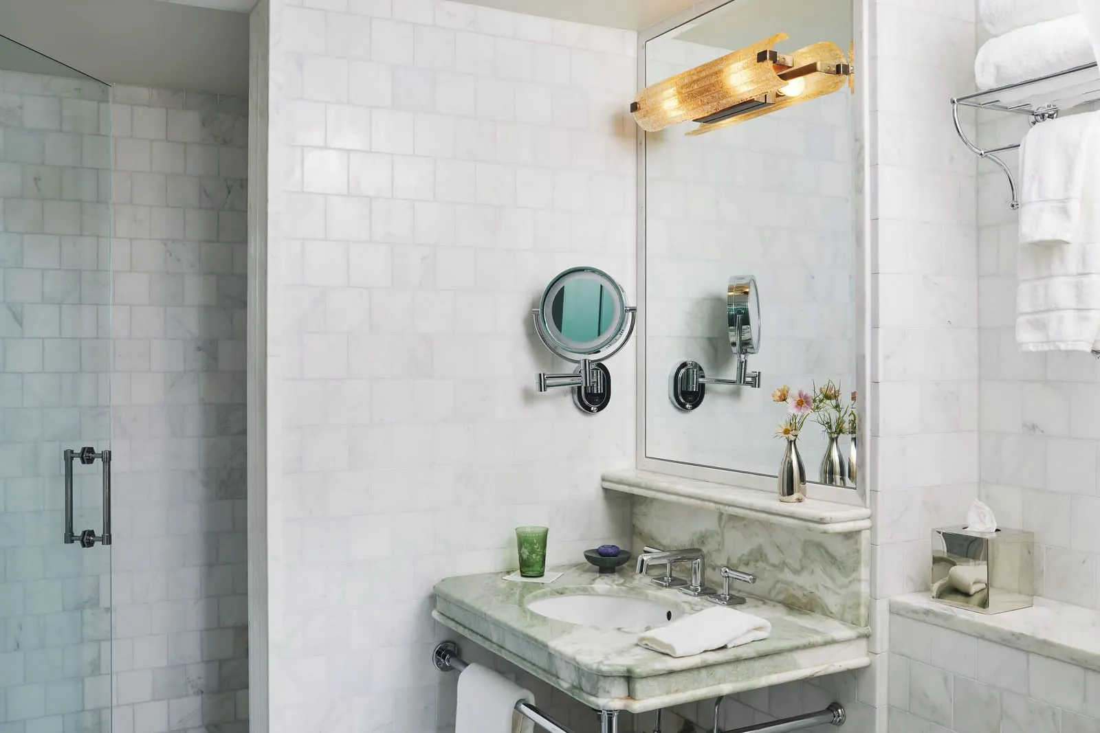 The Corner King at The Fifth Avenue Hotel: a marble bathroom with a glass doored shower, a green marble basin on a metal stand and a round magnifying mirror