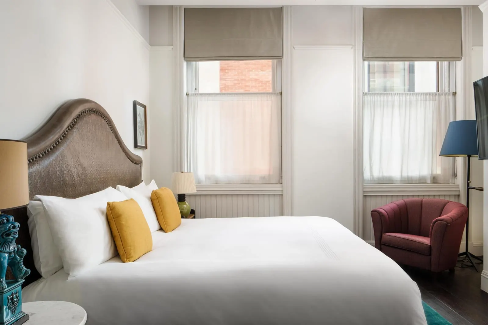 One Bedroom Corner Suite at The Beekman: a large bed with a studded leather headboard under two tall windows with grey blinds, and a burgundy armchair in the corner