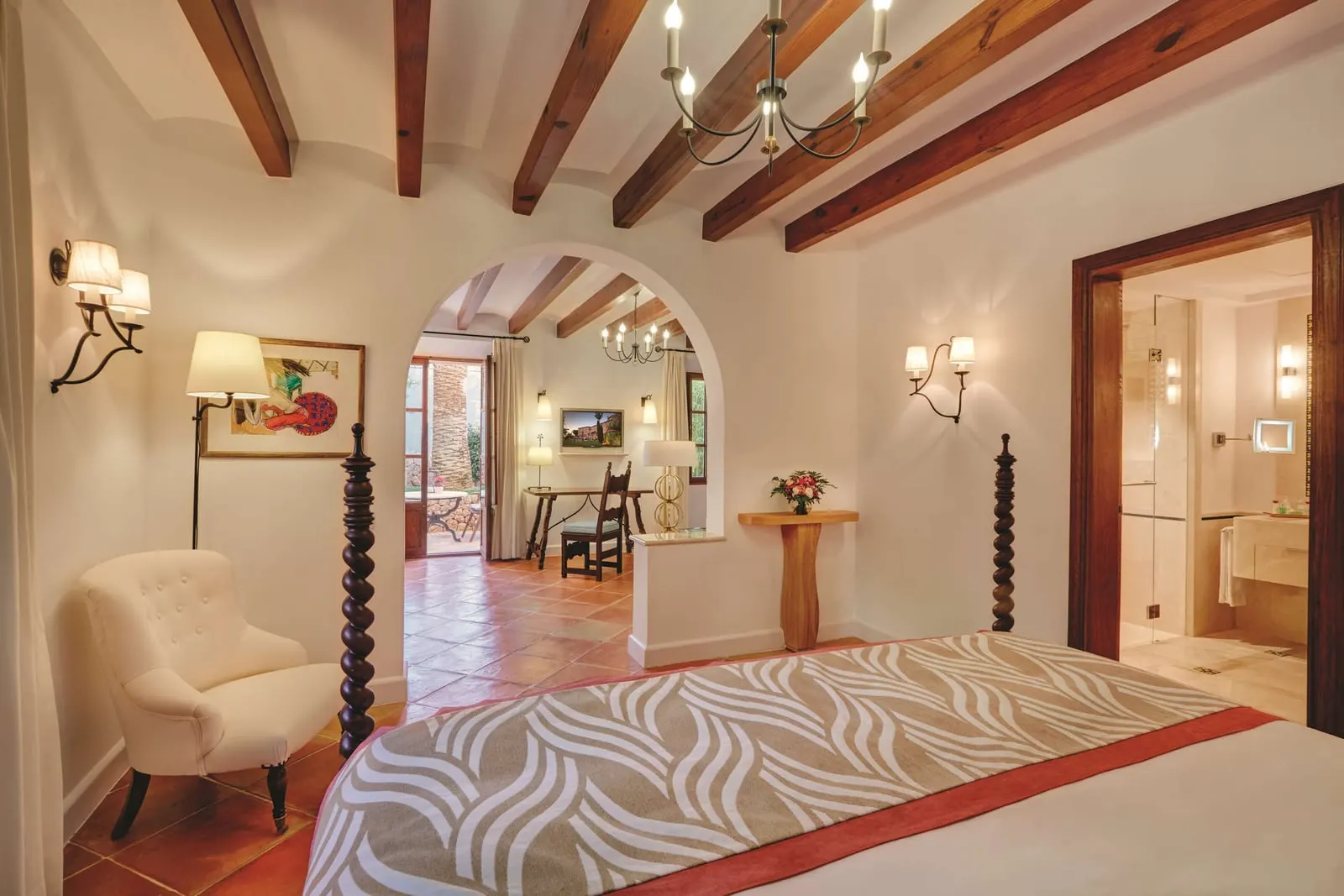 Junior Suite at Belmond La Residencia: a four-poster bed under wooden beams, looking through an arch to a desk and a tiled sitting area, with the bathroom door on the right