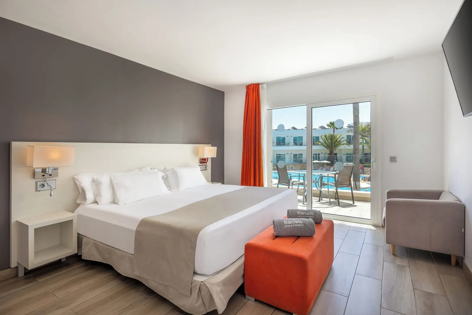 Suite Pool View at Barceló Corralejo Sands: a large bed with an orange footstool, an armchair and glass doors onto a terrace over the pool.