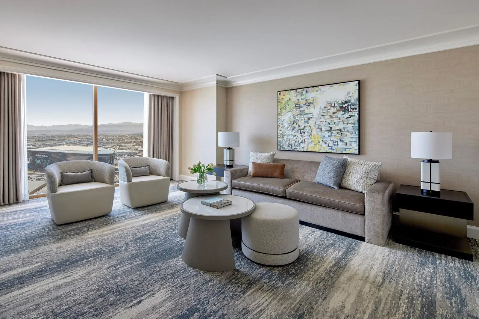 Stadium View Executive Suite at Four Seasons Hotel Las Vegas: a living room with a sofa, round tables and two armchairs by a floor-to-ceiling window onto Allegiant Stadium and the valley