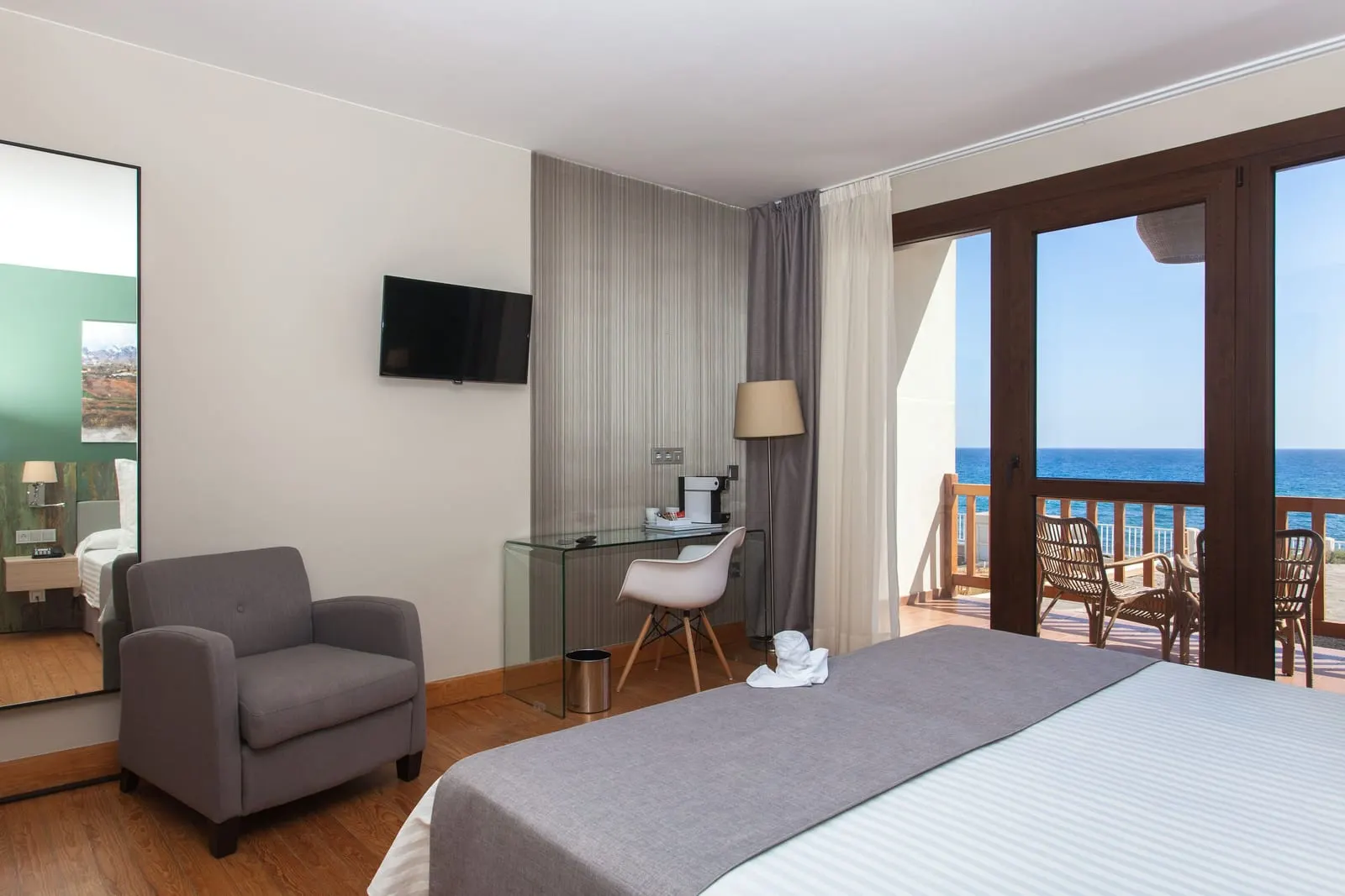Sea View Double Room at El Mirador de Fuerteventura: an armchair, glass desk and wall television beside doors to a balcony with two wicker chairs and the sea beyond