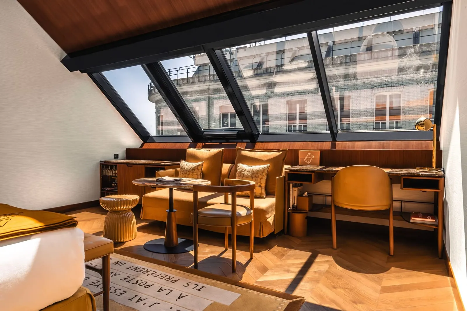 Junior Suite Atelier at Hôtel Madame Rêve: a sofa, a round table, an armchair and a writing desk beneath a wall of sloping atelier windows, facing the stone facade and iron balconies of the building opposite
