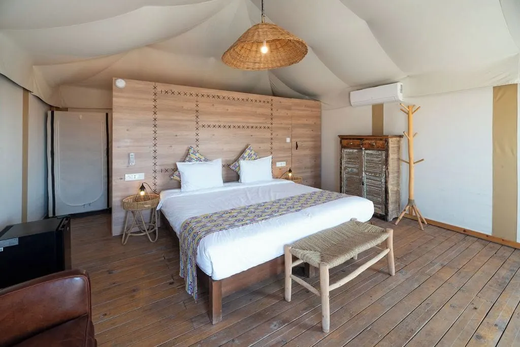 Emotion Tent at Inara Camp: a double bed with a patterned runner and a wooden bench under a woven pendant lamp and the white tent roof.