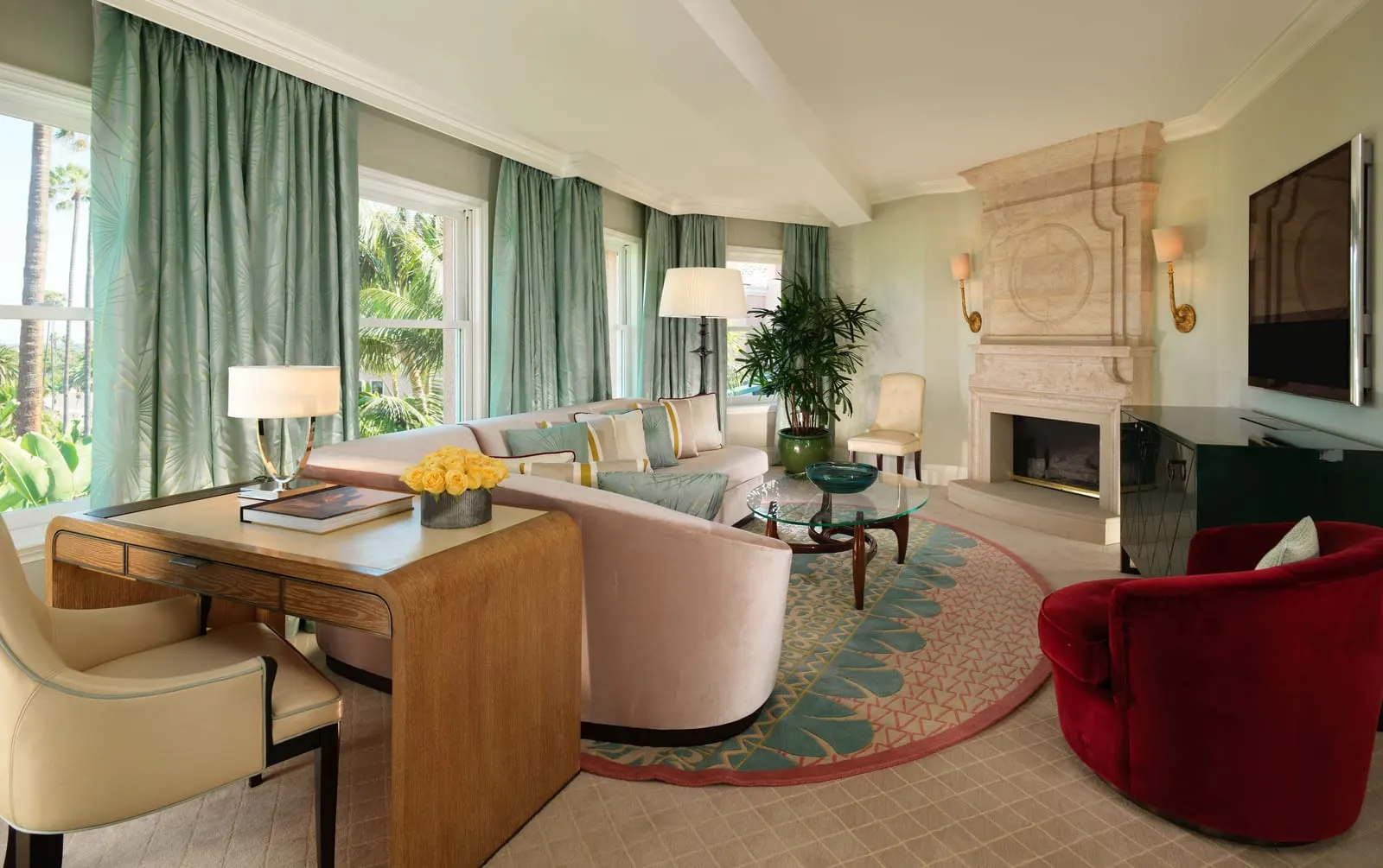 Beverly Hills Suite at The Beverly Hills Hotel: a living room with a curved pale pink sofa, a red velvet armchair and a glass coffee table on a round rug in front of a stone fireplace, with pale green curtains at the windows