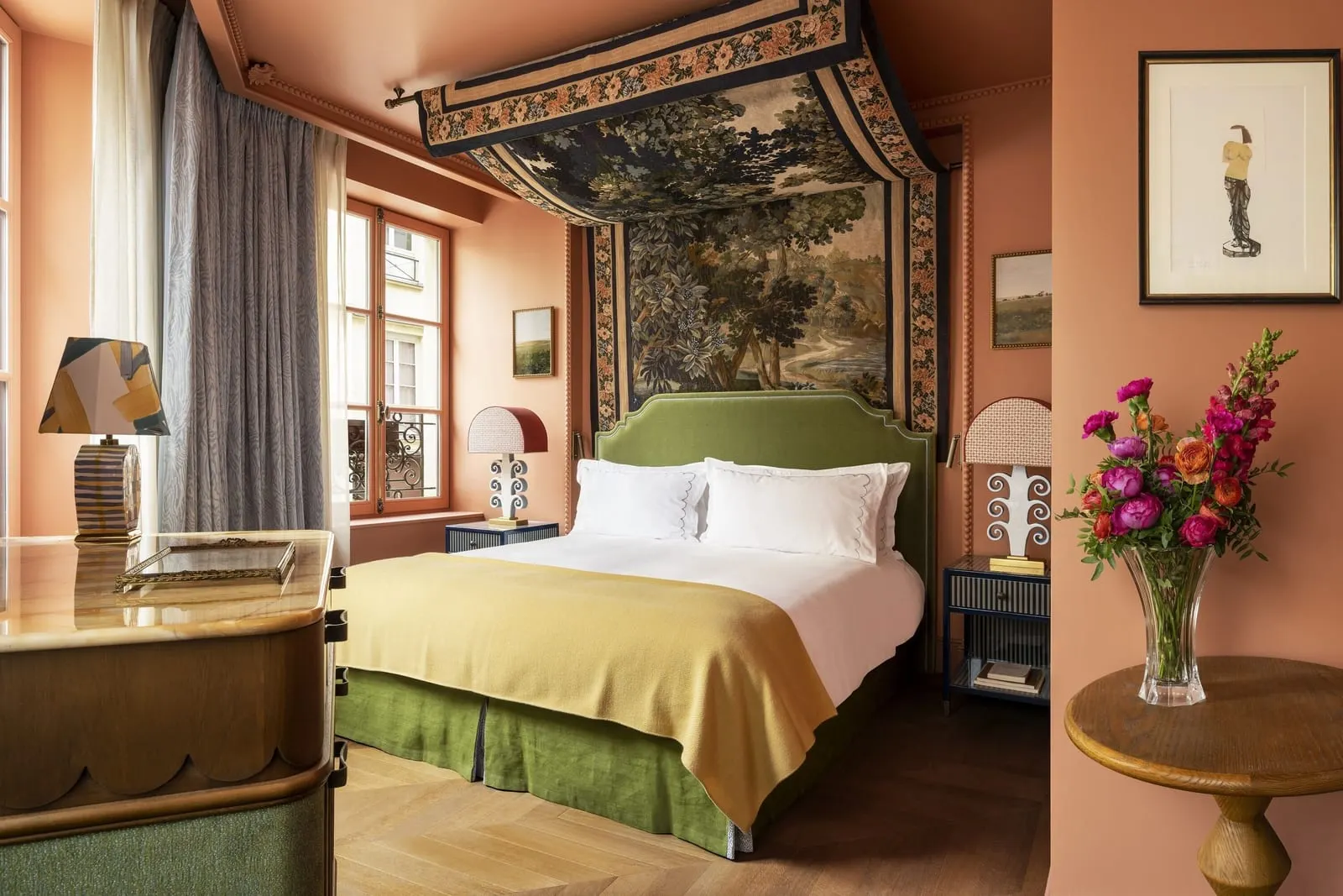 Deluxe Room at Le Grand Mazarin: a bed with a green velvet headboard and valance under a tapestry canopy, terracotta walls hung with framed pictures and a vase of bright flowers on a side table