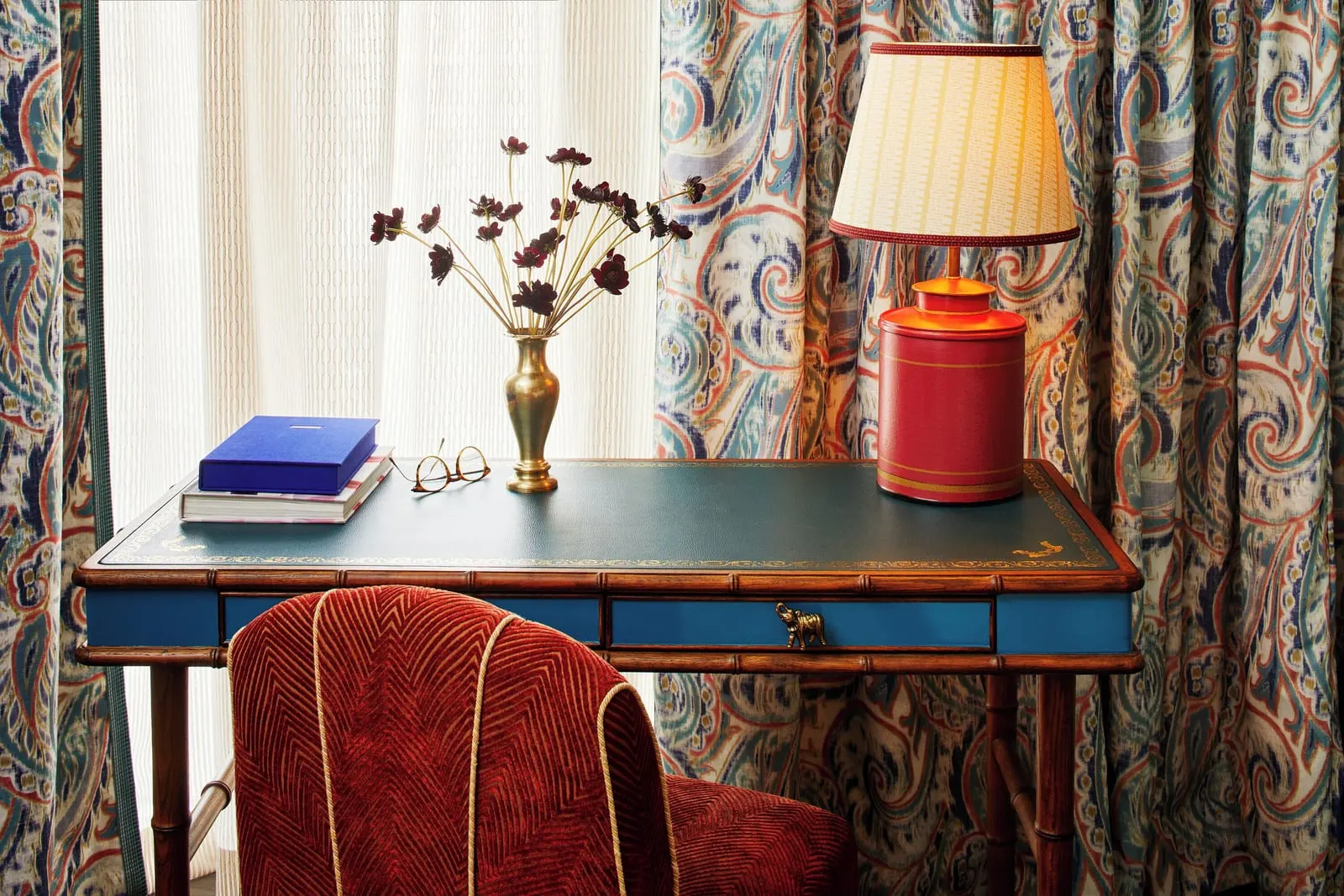 The Corner King at The Fifth Avenue Hotel: a leather topped writing desk with a brass vase of dark flowers, books and a red lamp, in front of a window with paisley curtains