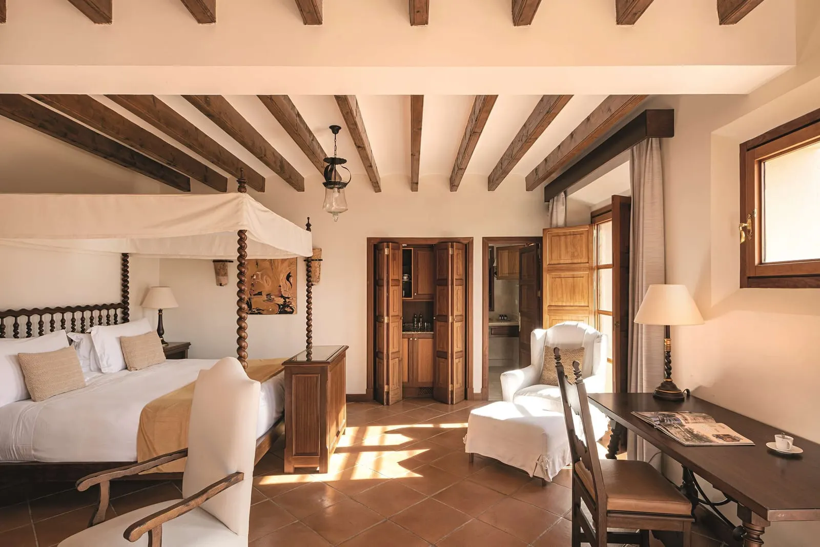 Executive Junior Suite at Belmond La Residencia: a four-poster bed with a white canopy under wooden beams, a white armchair with a footstool, a desk and folding wooden doors, on a terracotta tiled floor