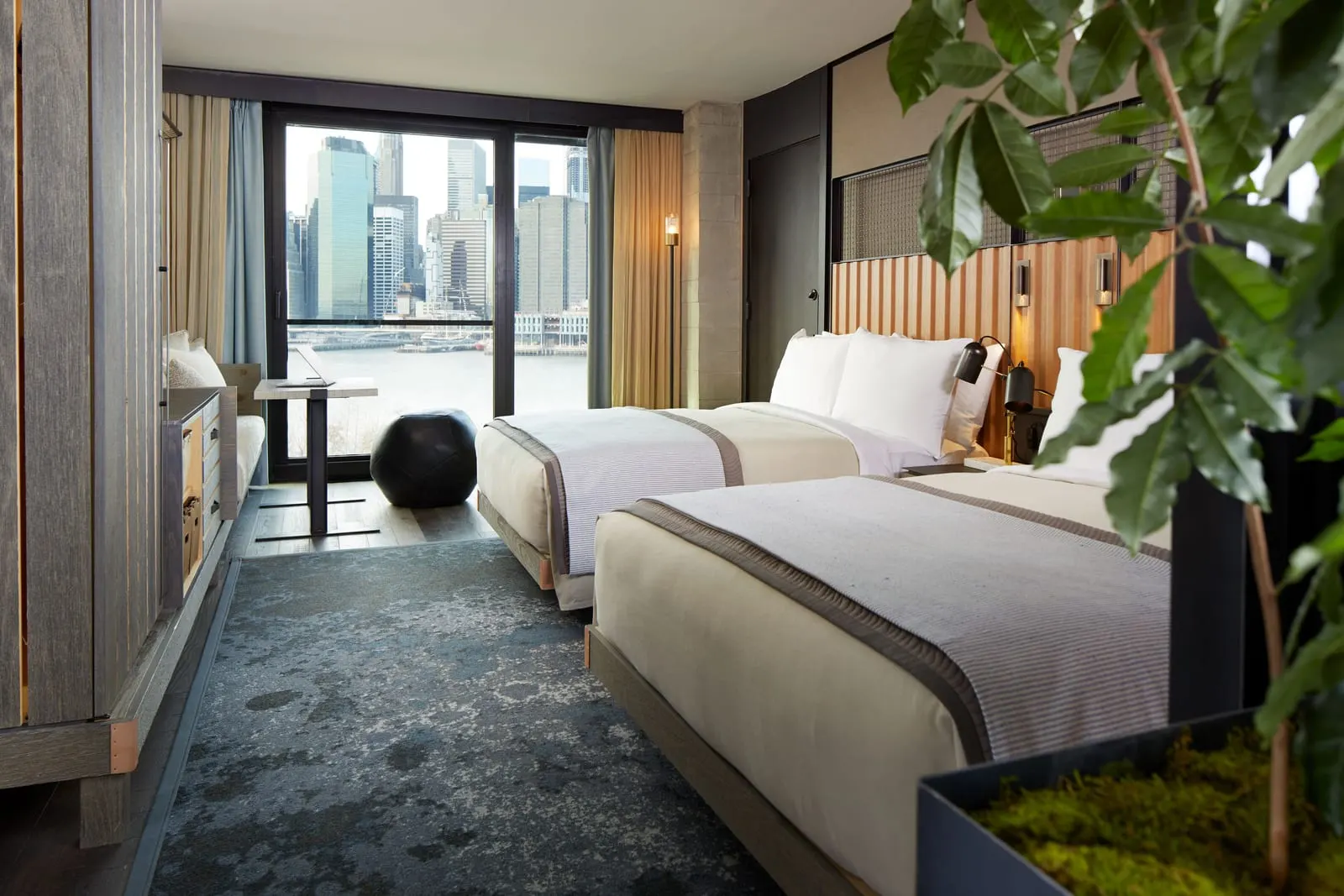 Harbor 2 Beds im 1 Hotel Brooklyn Bridge: zwei Doppelbetten gegenüber einem raumhohen Fenster zum East River, mit einem Schreibtisch, einem runden schwarzen Pouf und hohen Gebäuden am gegenüberliegenden Ufer