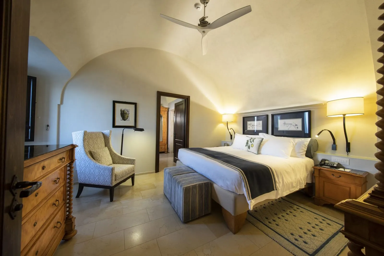 Premium Terrace Suite at Monastero Santa Rosa: a king bed under a vaulted ceiling with a ceiling fan, an armchair and a wooden chest of drawers.