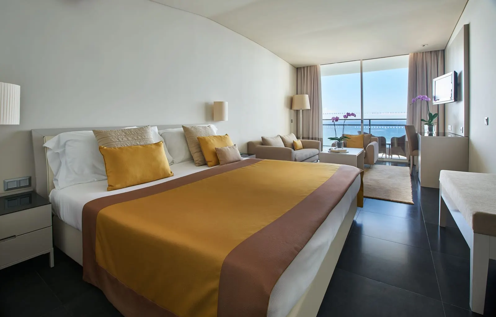 VidaMar Resort Hotel Madeira Premium Sea View：マスタード色のクッションのキングサイズのベッドと、ソファ、バルコニーと海に面したガラスの壁
