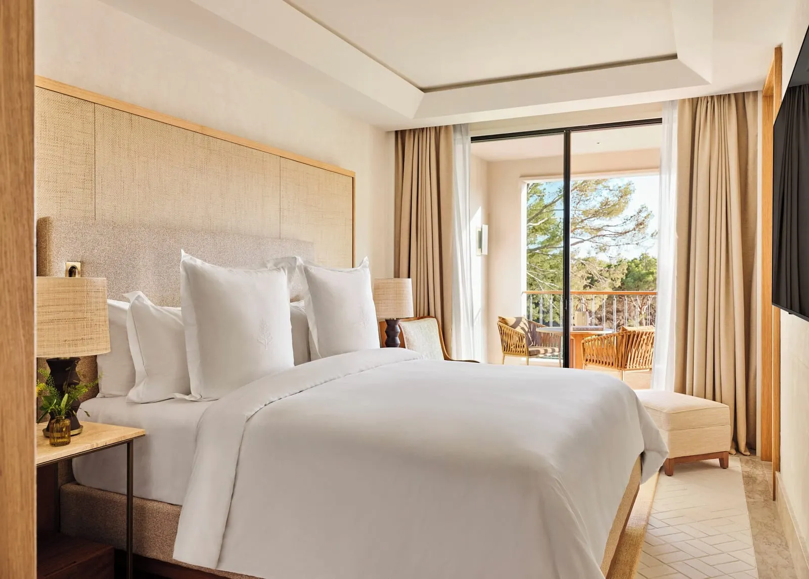 Pine-View Suite im Four Seasons Resort Mallorca at Formentor: ein Kingsize-Bett mit einem geflochtenen Kopfteil und einem Wandfernseher, mit einer Glastür zu einer Terrasse mit Stühlen und Kiefern