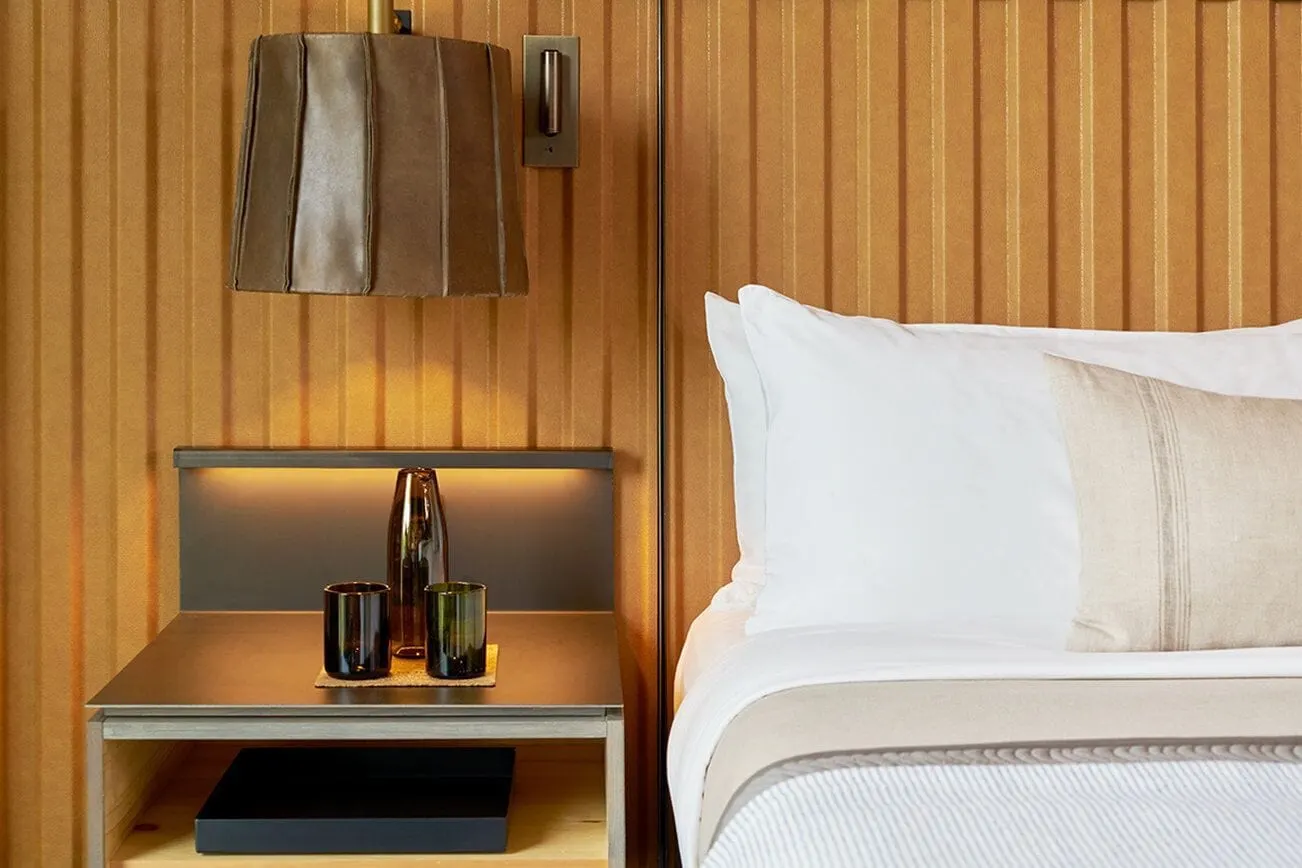 Bridge King im 1 Hotel Brooklyn Bridge: ein Nachttisch mit zwei grünen Gläsern und einer Karaffe unter einer Wandlampe mit Lederschirm, neben einem Kingsize-Bett mit weißen Kissen und einem Leinenkissen