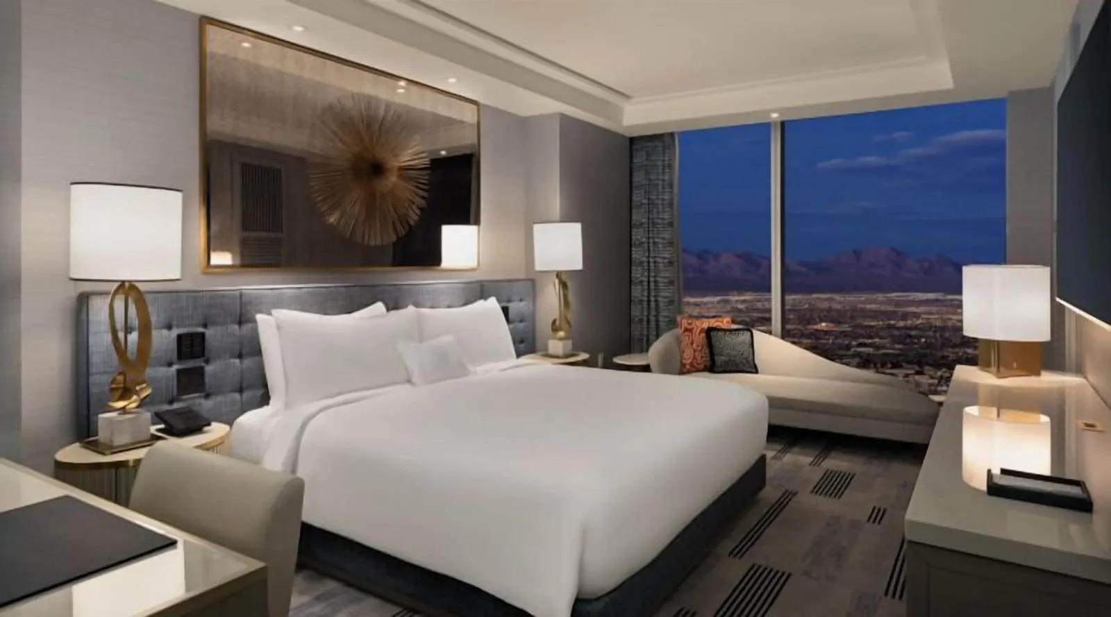 Regal Suite Mountain Great View at Fontainebleau Las Vegas: a king bed under a gilt-framed mirror with a gold starburst, and a chaise longue by a window onto the city lights and the mountains