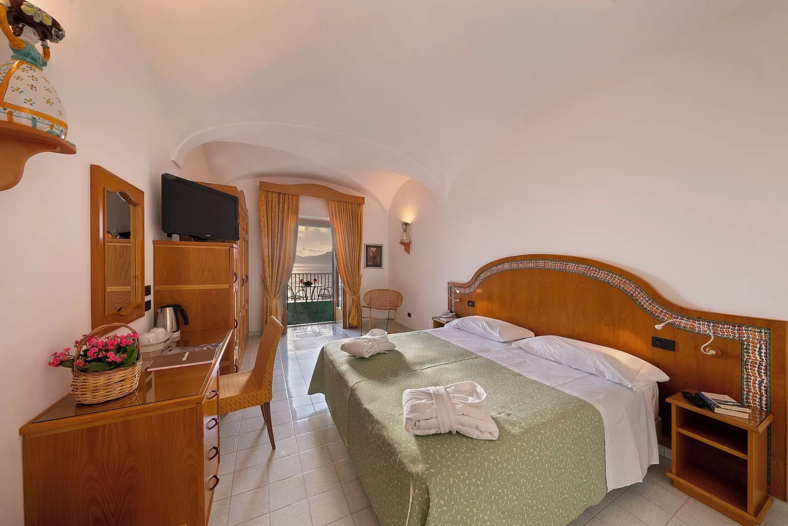 Classic Room with sea view terrace at Hotel Tramonto d'Oro: a double bed with a wooden headboard edged in ceramic tiles under a vaulted ceiling, with a wooden desk and wardrobe and curtained doors open to the terrace.