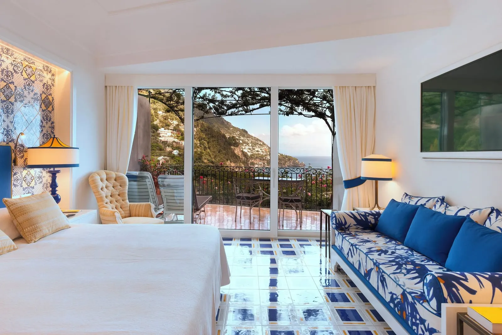 Elite Room at Il San Pietro di Positano: a double bed and a long blue sofa, with sliding doors to a vine-shaded terrace with a sunbed above the sea