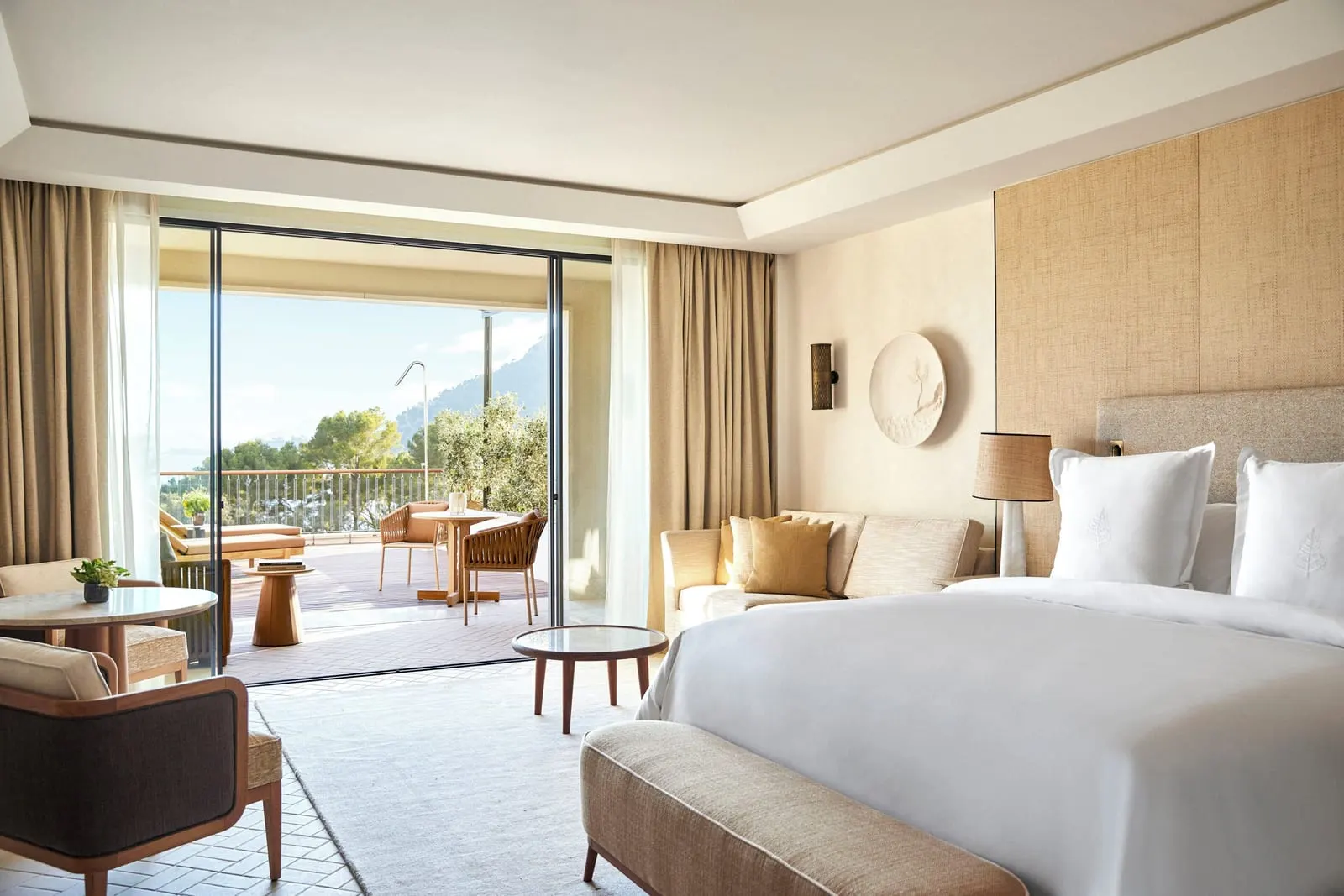 Grand Pine-View Room im Four Seasons Resort Mallorca at Formentor: ein Kingsize-Bett mit einer gepolsterten Bank am Fußende, ein Sofa und ein Sessel und Glastüren zu einer Terrasse mit Tisch und Stühlen