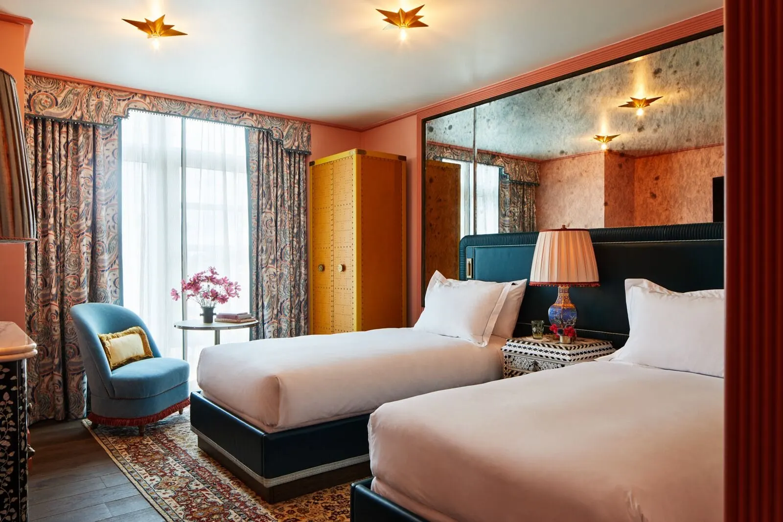 The NoMad Twin at The Fifth Avenue Hotel: two single beds with white linen and dark blue bases, a studded wardrobe and a blue armchair by a tall window, with a mirrored wall behind the beds