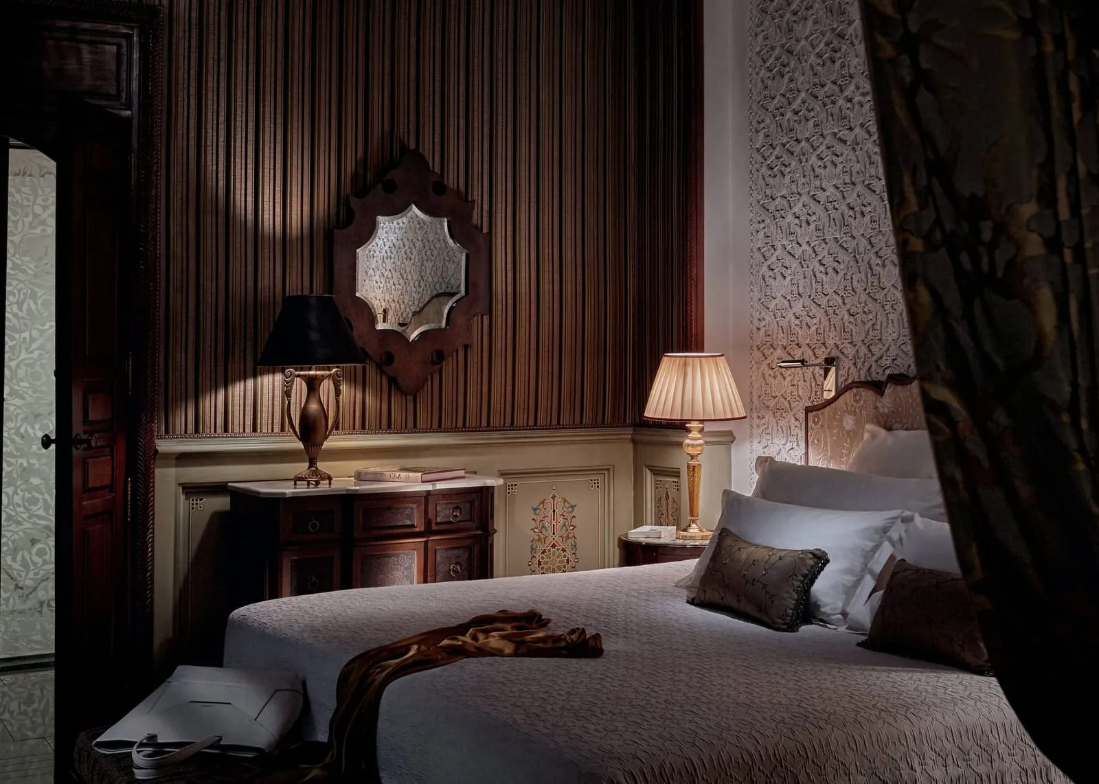 Privilege Riad at Royal Mansour Marrakech: a bed with grey cushions beside a carved chest of drawers, two table lamps and an ornate mirror on a striped wall