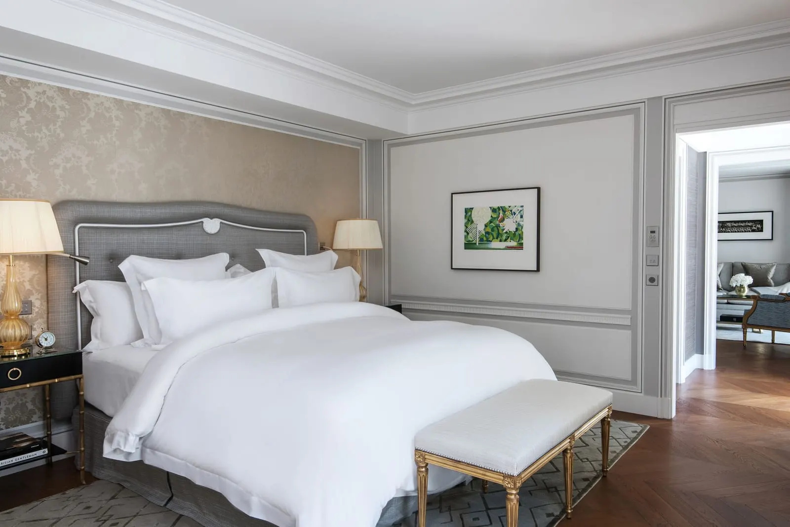 Premier Suite at Hôtel de Crillon, A Rosewood Hotel: a king bed with a grey upholstered headboard against damask wallpaper, a gilt bench at its foot and the lounge through an open doorway