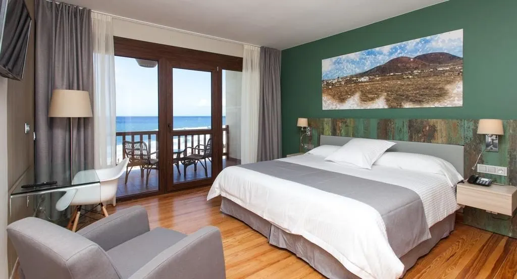 Sea View Double Room at El Mirador de Fuerteventura: a king bed below a painting of the island, with glass doors open to a balcony over the sea