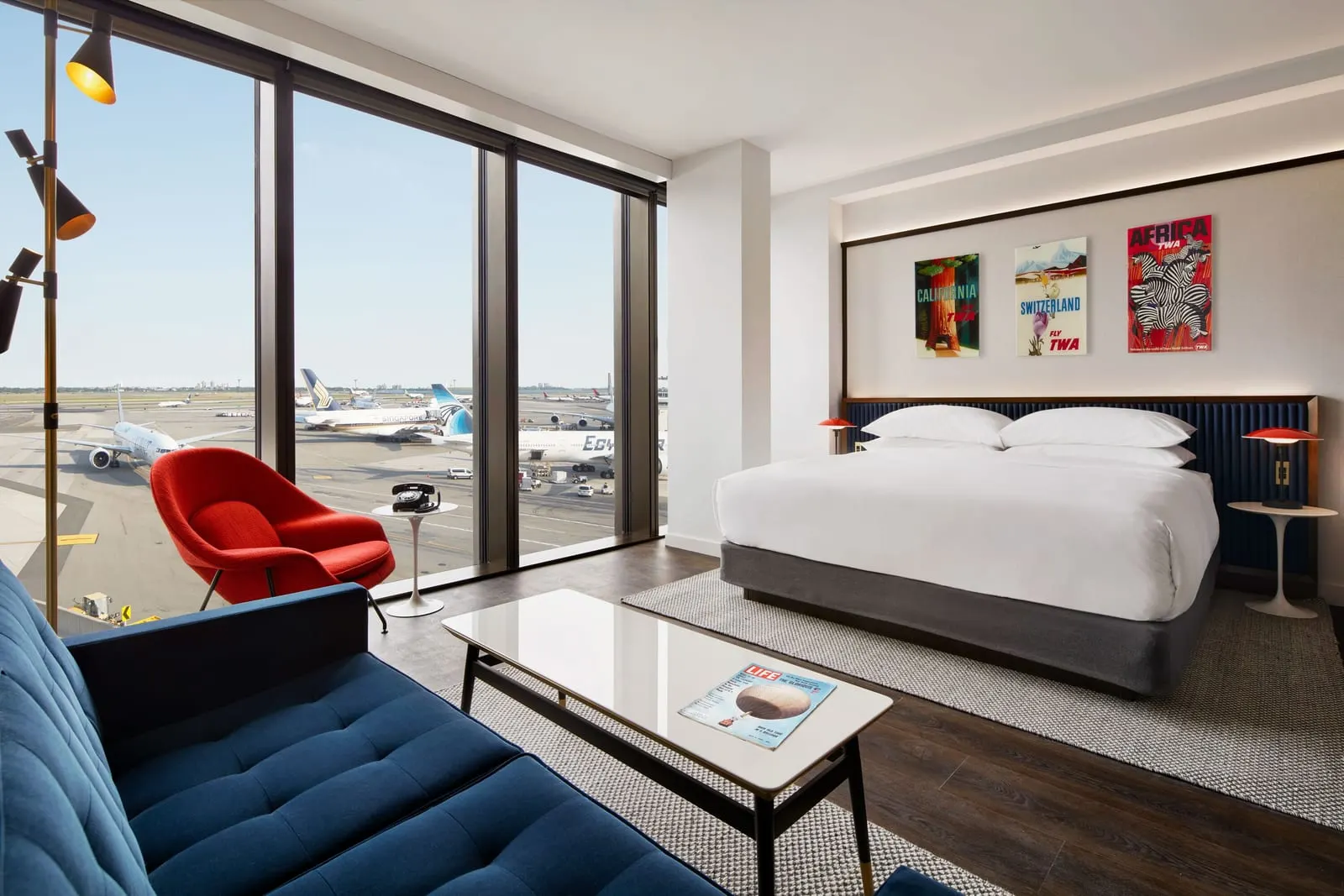 Executive King Suite With Runway View at TWA Hotel: a king bed, a blue sofa and a red armchair beside floor-to-ceiling windows overlooking airliners standing on the apron