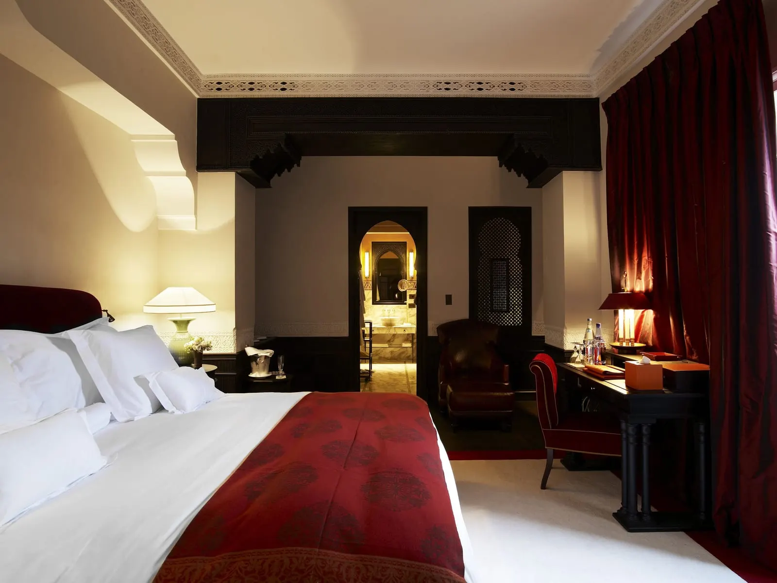 Hivernage Suite at La Mamounia: a king bed with a red throw, a desk and deep red curtains beneath a carved dark wood beam