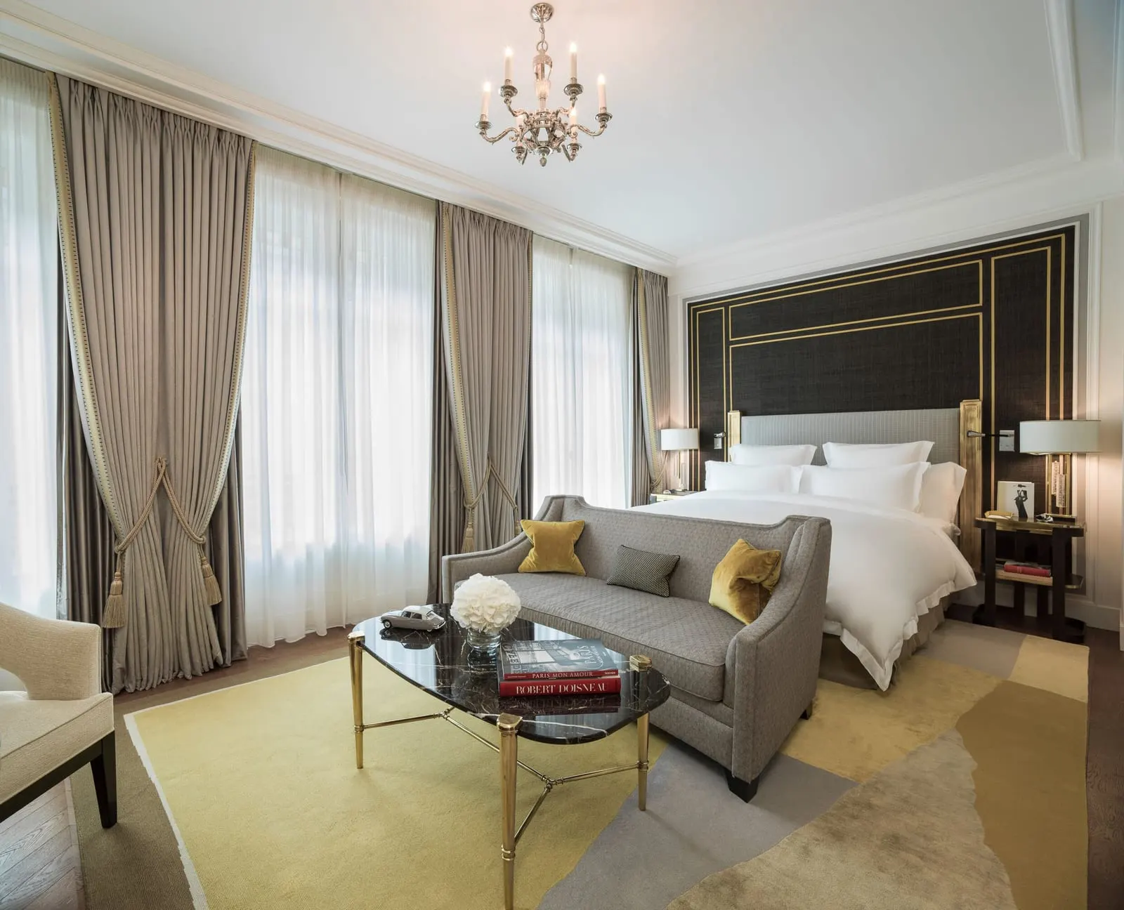 Junior Suite at Hôtel de Crillon, A Rosewood Hotel: a king bed against a dark wall lined in brass, a sofa and a marble coffee table at its foot, and a row of tall windows with sheer curtains under a chandelier